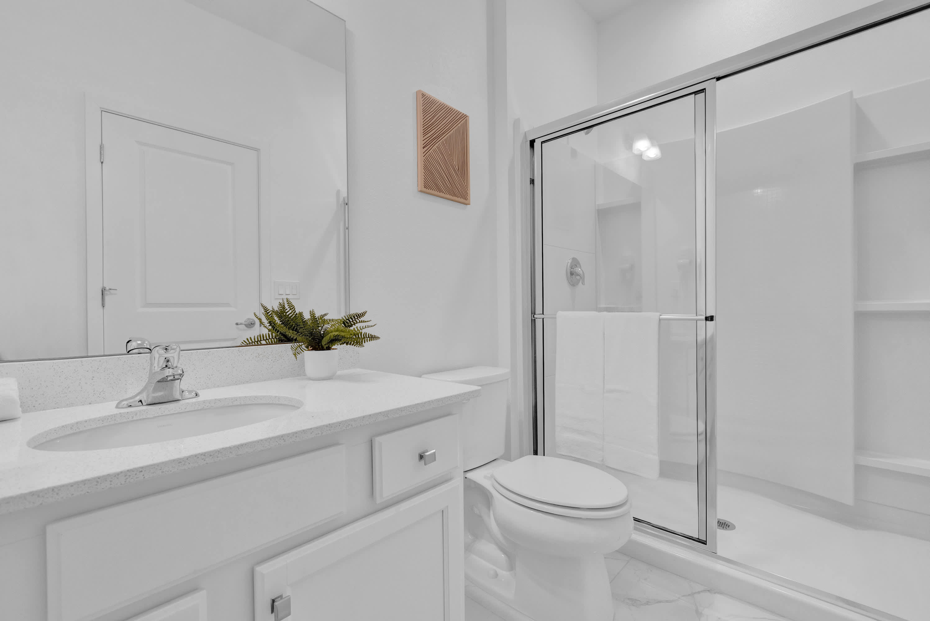 Clean white bathroom featuring a single vanity with a white quartz countertop, a toilet, and a glass-enclosed shower. A small potted plant sits on the vanity, and a textured wooden art piece hangs on the wall.