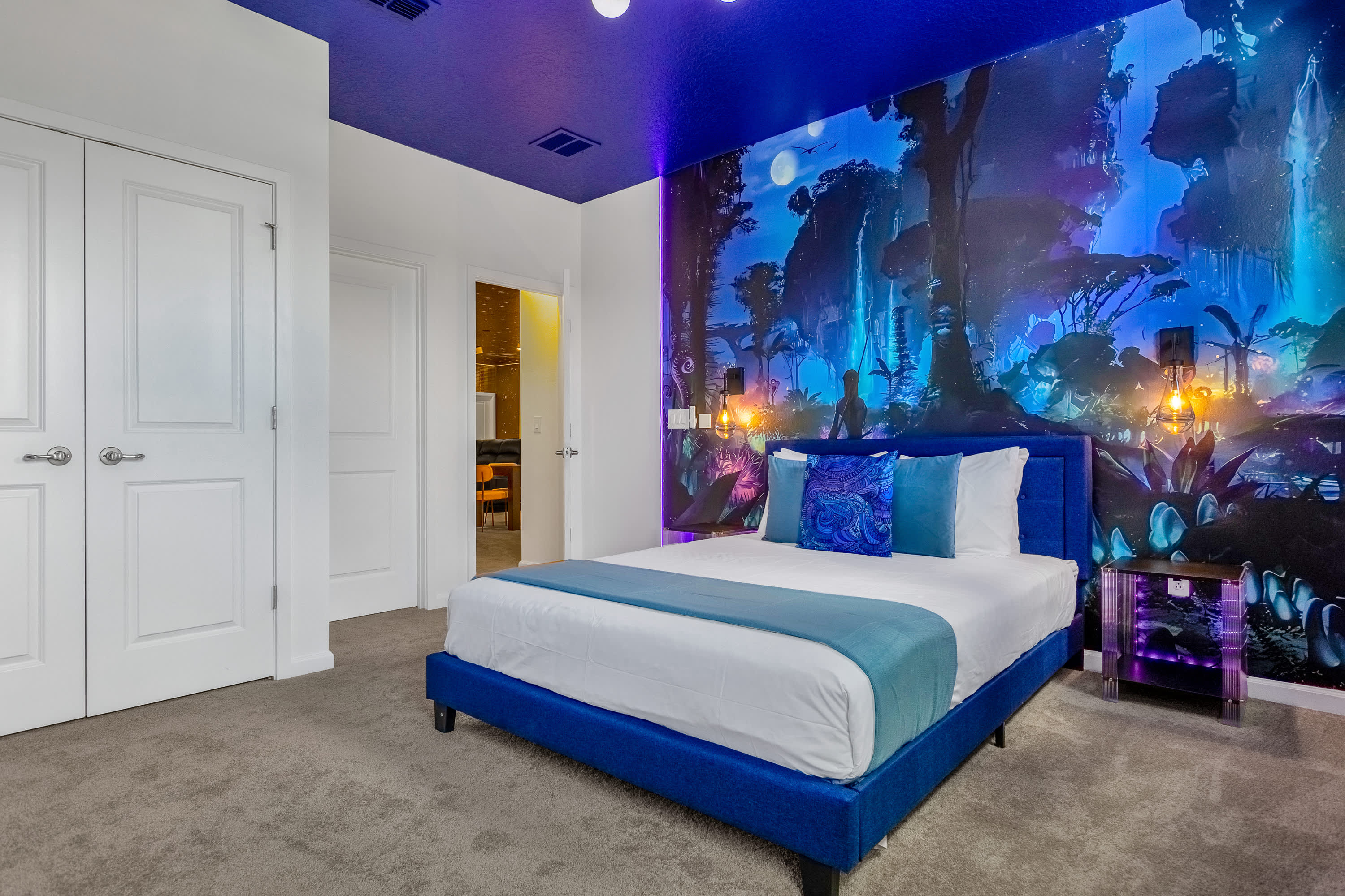 Another view of the vibrant, themed bedroom with a fantastical forest mural covering one wall and a deep blue ceiling. A blue upholstered bed with white bedding and blue pillows is visible, along with white double doors.