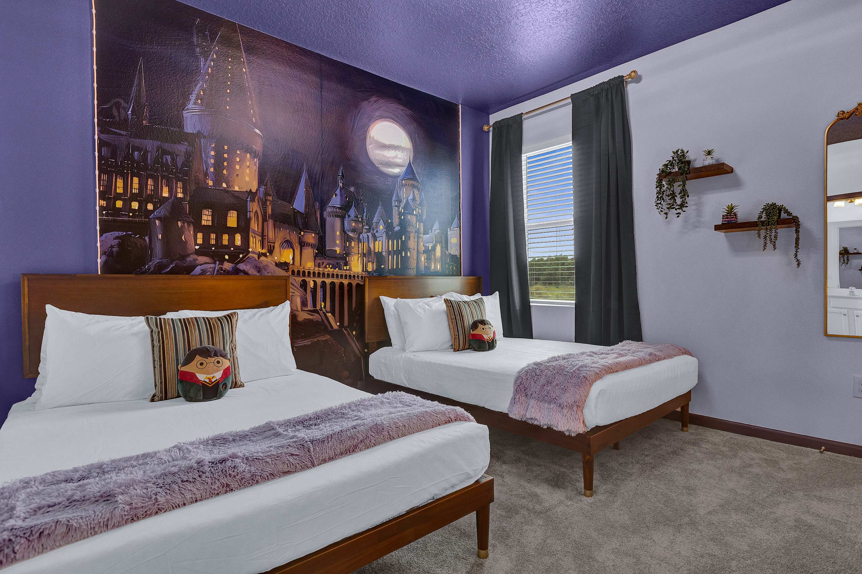 A Harry Potter themed bedroom featuring a large mural of Hogwarts castle on one wall and a dark purple ceiling. Two twin beds with wooden frames, white bedding, and purple throws are arranged side-by-side.