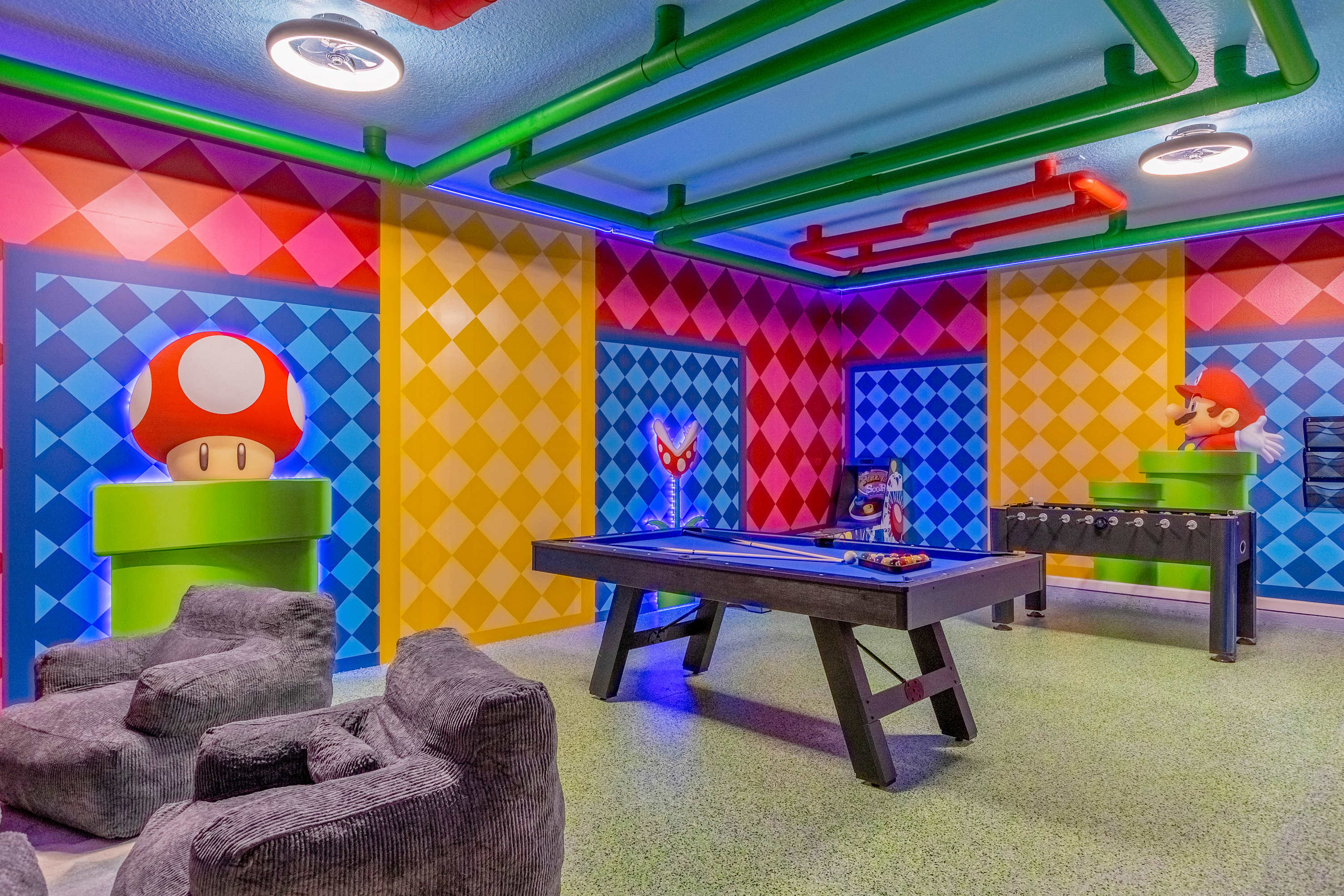 A vibrant Mario-themed game room featuring a pool table, foosball table, and comfortable bean bag chairs, all set against colorful checkered walls and illuminated by bright LED lighting.