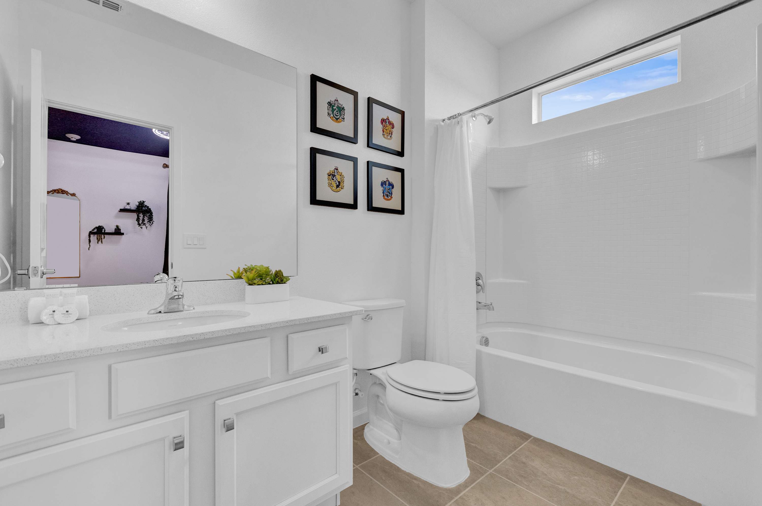 A clean white bathroom with a single vanity, a large mirror reflecting the Harry Potter themed bedroom, and a toilet. A white tiled tub and shower combo is on the right, with a horizontal window above it.