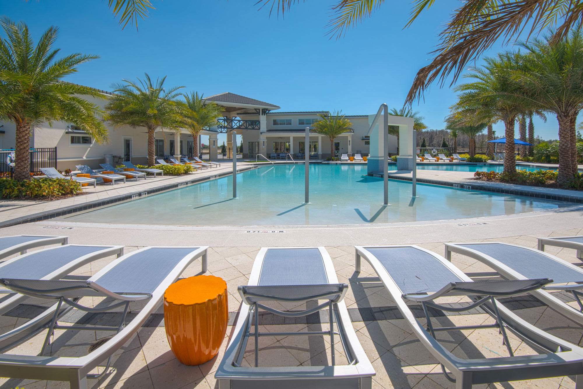 A large, resort-style outdoor swimming pool with a shallow entry area, surrounded by numerous lounge chairs on a light-colored paved deck. Palm trees and a modern clubhouse building frame the sunny scene under a clear blue sky.