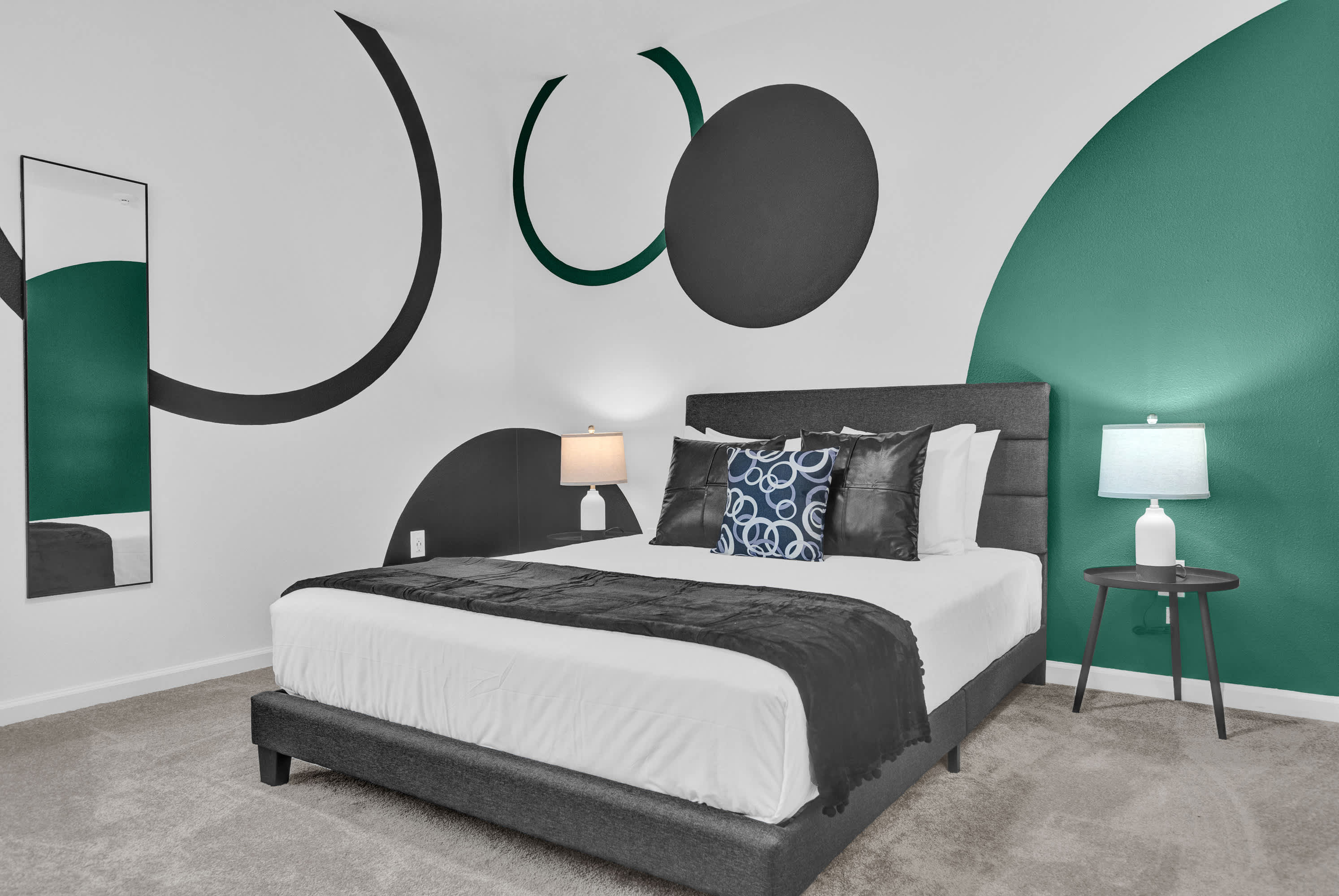 Contemporary bedroom with a striking green and black geometric mural on the wall, a king bed with a black upholstered headboard, and a black nightstand with a white lamp. A full-length mirror is visible on the left.