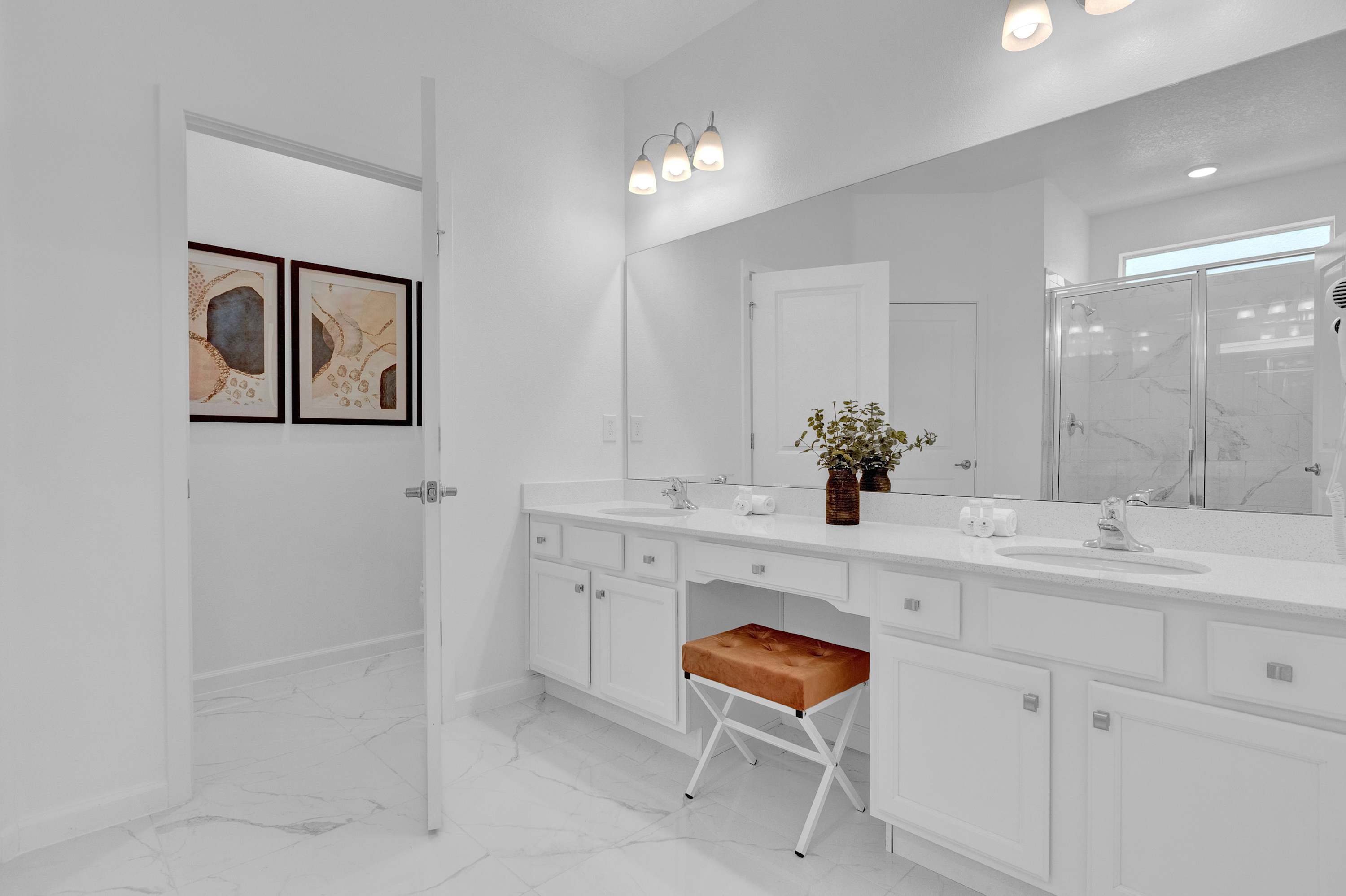 A bright, modern bathroom featuring a double vanity with white quartz countertops, dual sinks, and a large mirror, complemented by a burnt orange upholstered stool and white marble-look floor tiles.