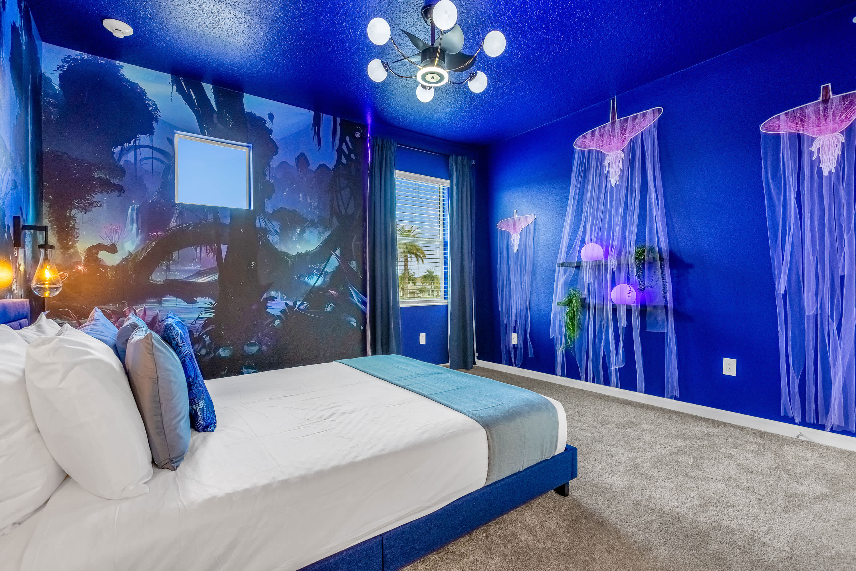 A vibrant, themed bedroom with a deep blue ceiling and a large, colorful mural depicting a fantastical forest scene. A blue upholstered bed with white bedding and blue pillows is centered in the room.