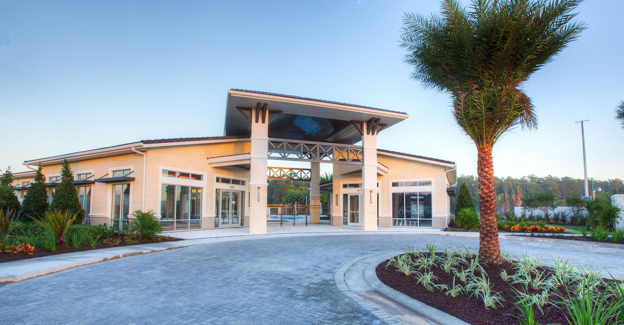 The grand exterior entrance of a modern clubhouse building features a distinctive architectural roofline and large windows, surrounded by lush landscaping. A circular paved driveway leads up to the welcoming facade under a clear sky.