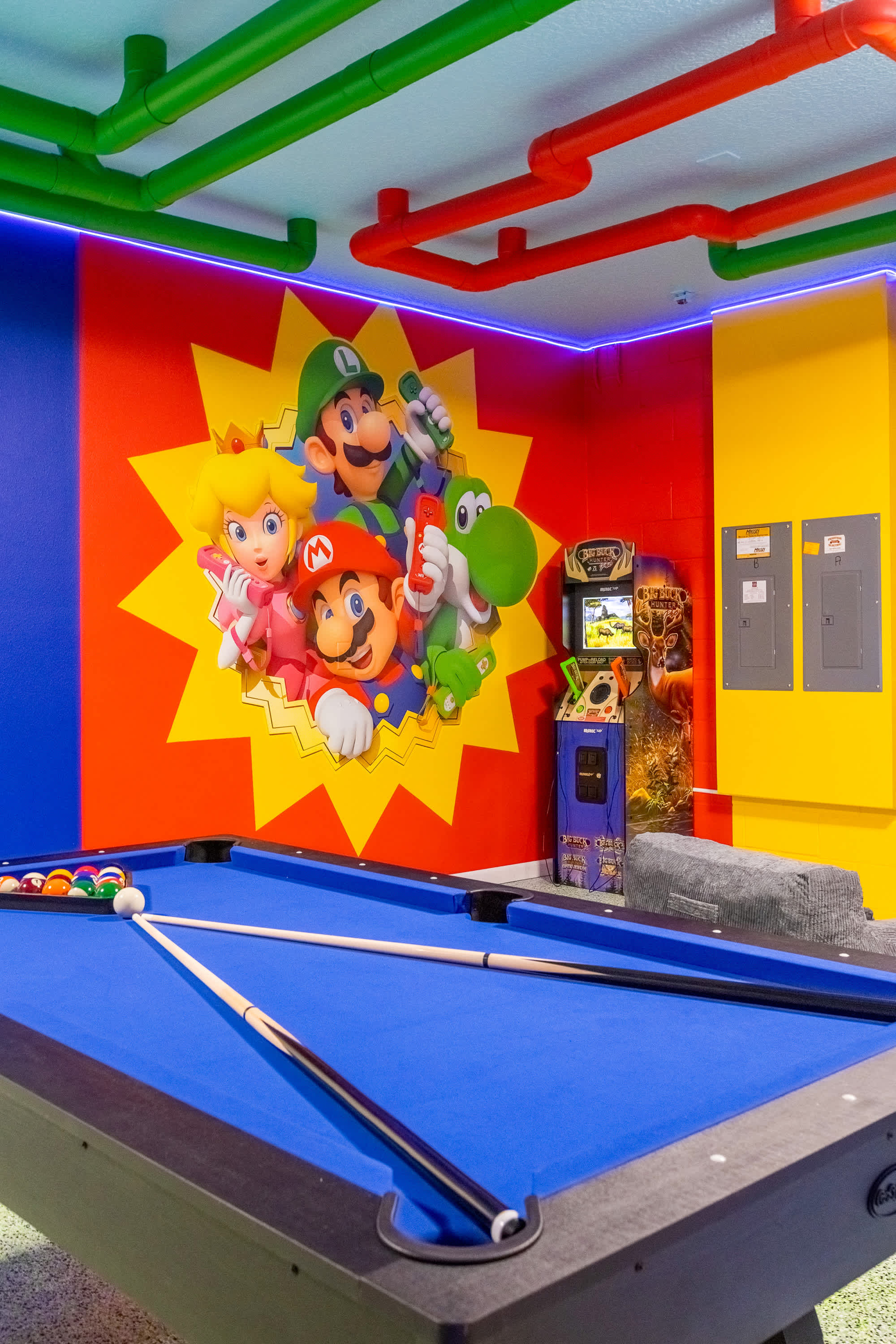 Vibrant game room featuring a blue felt pool table with cues and balls, set against a red wall with a large Mario Bros. mural, illuminated by blue LED strip lighting and colorful overhead pipes.