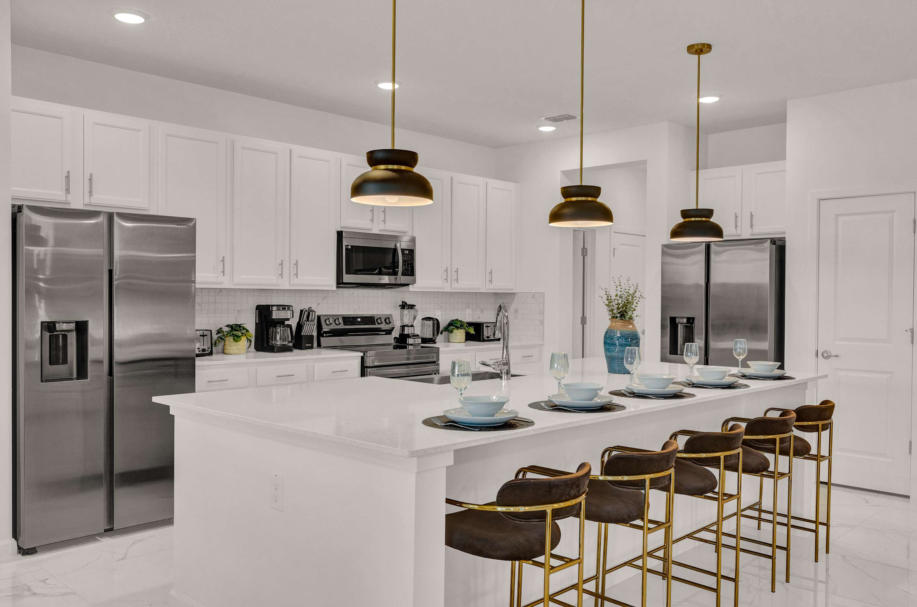 Bright, modern kitchen with white shaker cabinets, stainless steel appliances, and a large island with white countertops and four bar stools. Gold pendant lights hang above the island, which is set with place settings.
