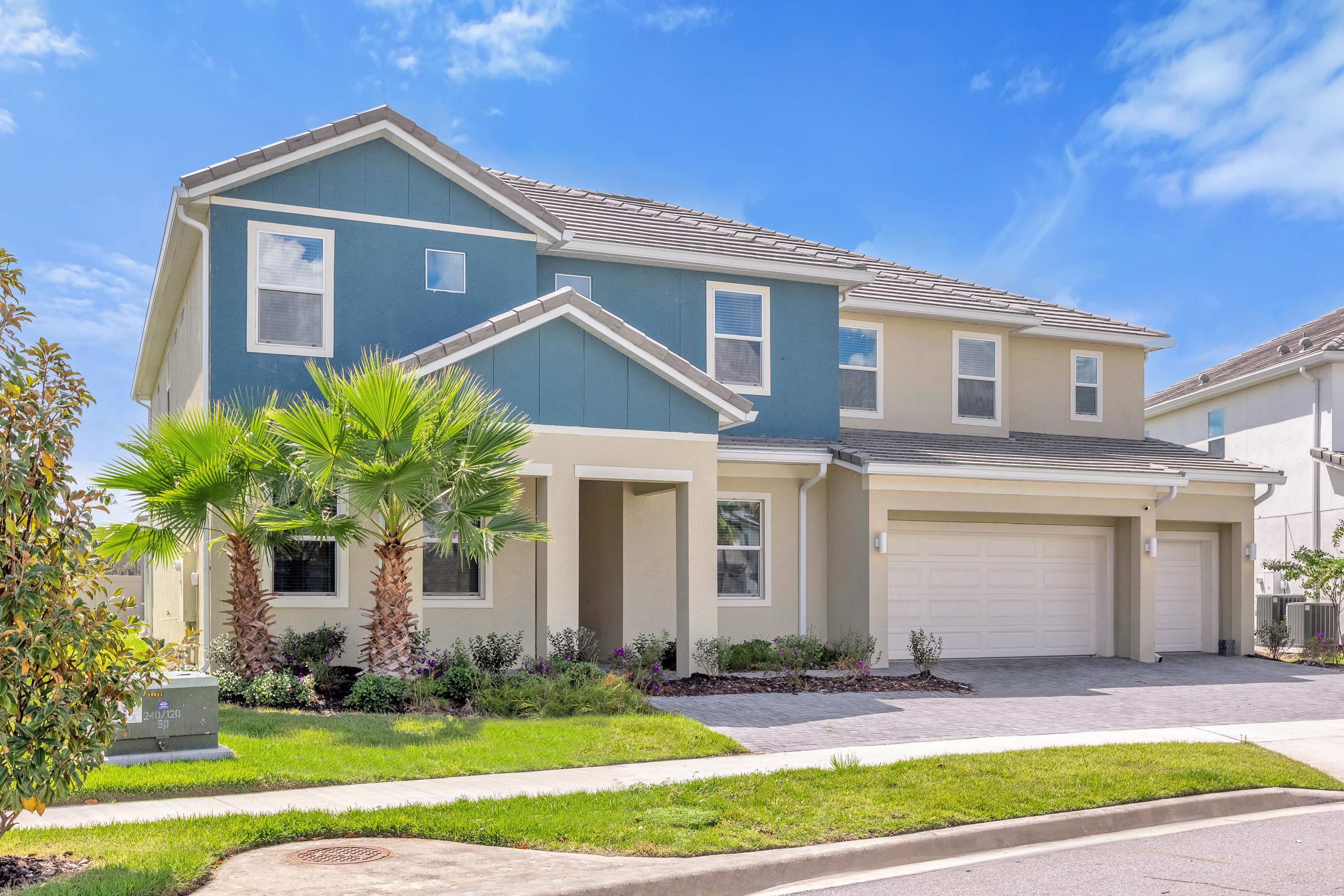 The front exterior of a large, two-story house with a blue and beige facade, featuring multiple windows and a two-car garage. Palm trees and landscaped shrubbery adorn the front yard, with a paved driveway leading to the garage.