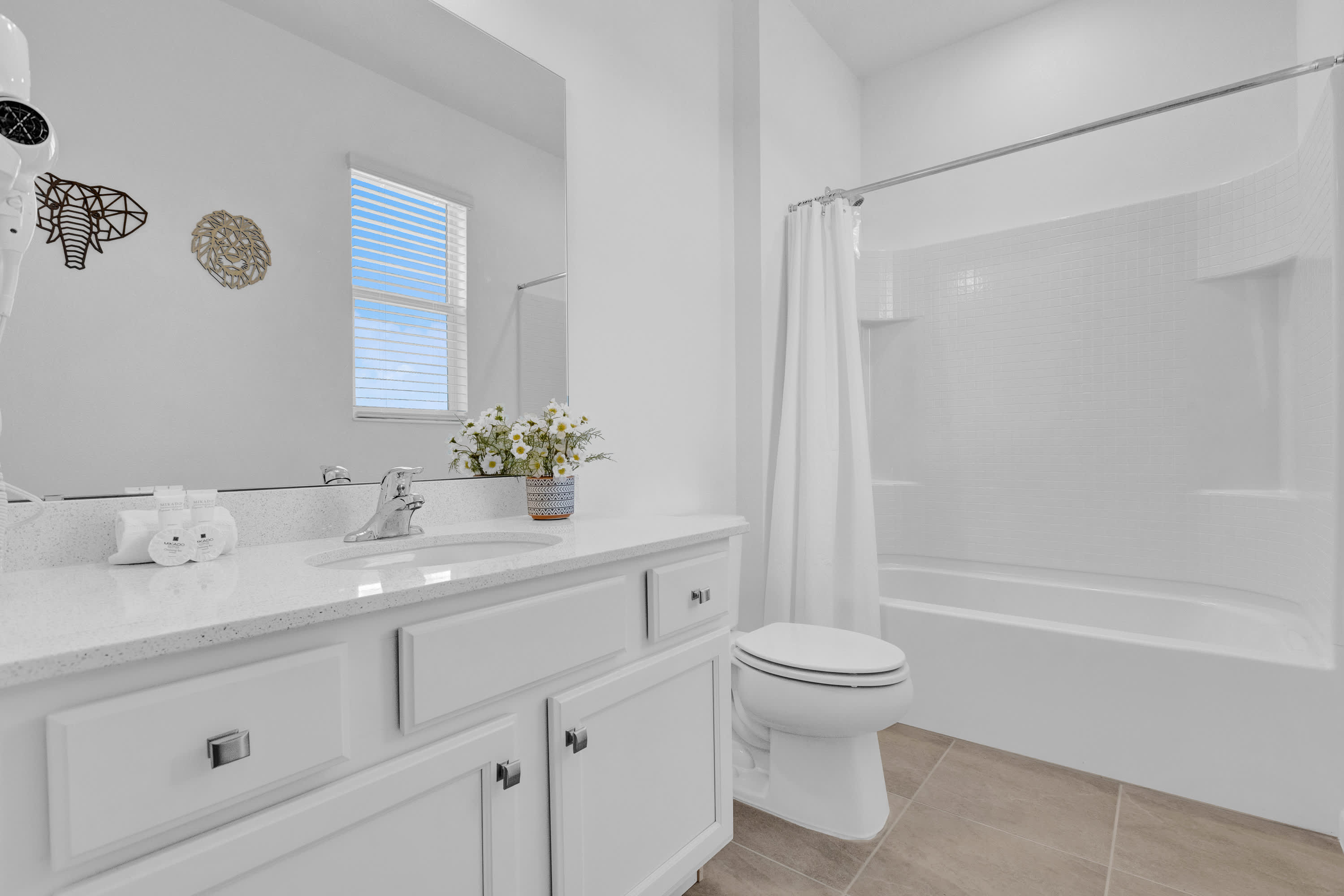 A bright and minimalist bathroom with a single white vanity, a large mirror, a toilet, and a bathtub with a shower curtain, featuring a small window with blinds and a decorative plant.