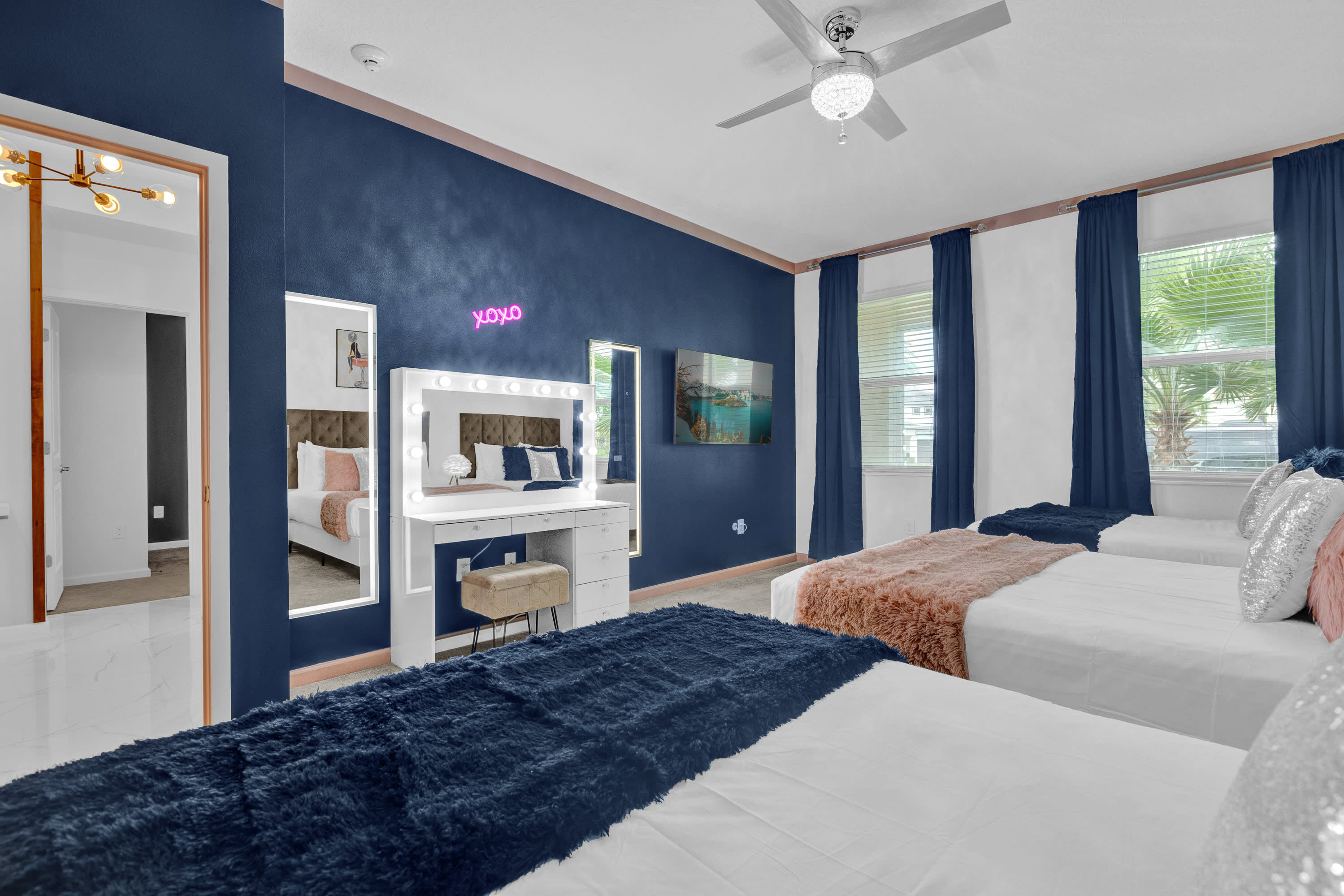 A vibrant bedroom with a dark blue accent wall featuring an "XOXO" neon sign, three beds with white bedding and plush blue and pink throws, and a white vanity with a lighted mirror.