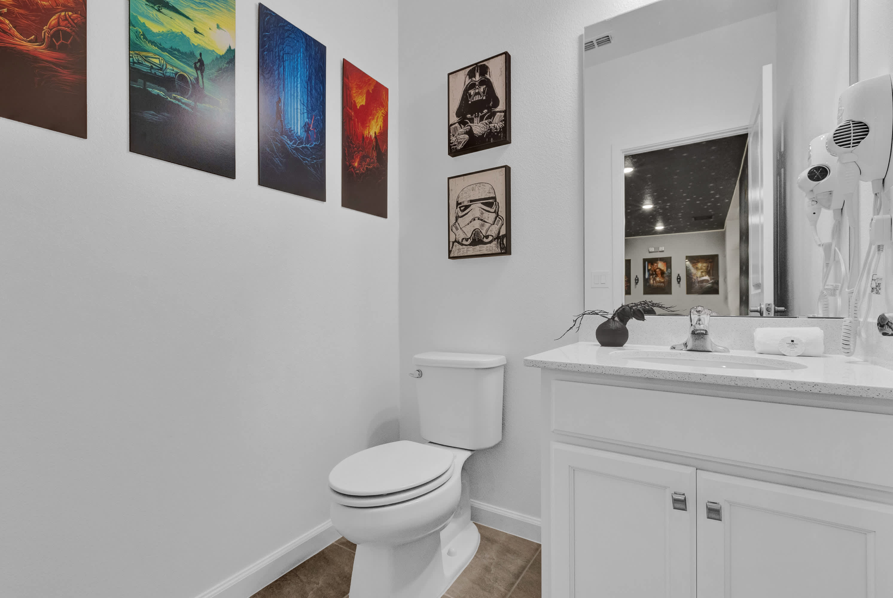 A modern bathroom featuring a white toilet and vanity with a white countertop and sink, complemented by Star Wars-themed artwork on the light gray walls. A large mirror reflects a dark, possibly star-patterned ceiling, and a hair dryer is mounted on the wall.