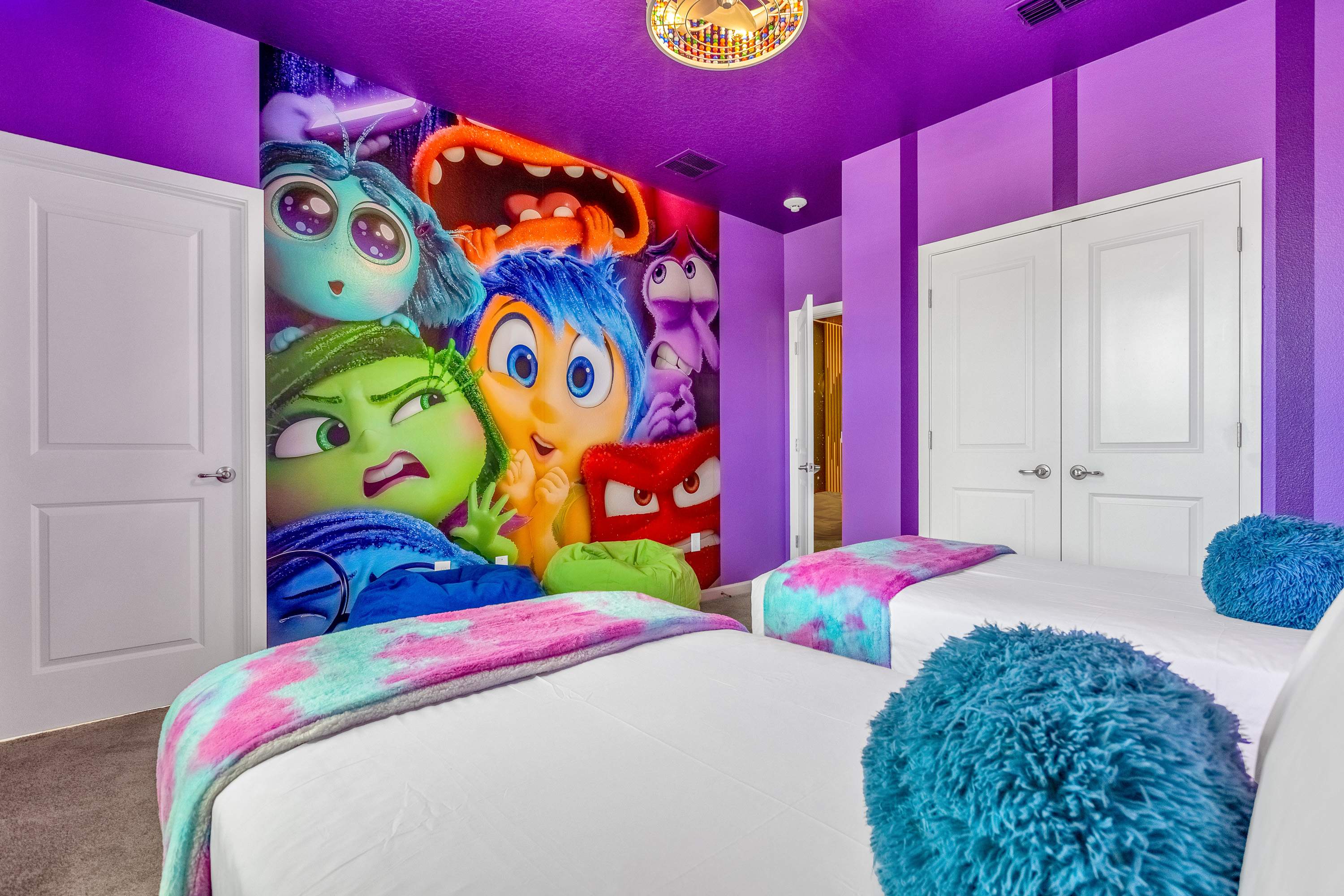 A whimsical bedroom with vibrant purple walls and ceiling, featuring a large "Inside Out" character mural, two beds with white bedding and colorful tie-dye throws, and a decorative ceiling light.