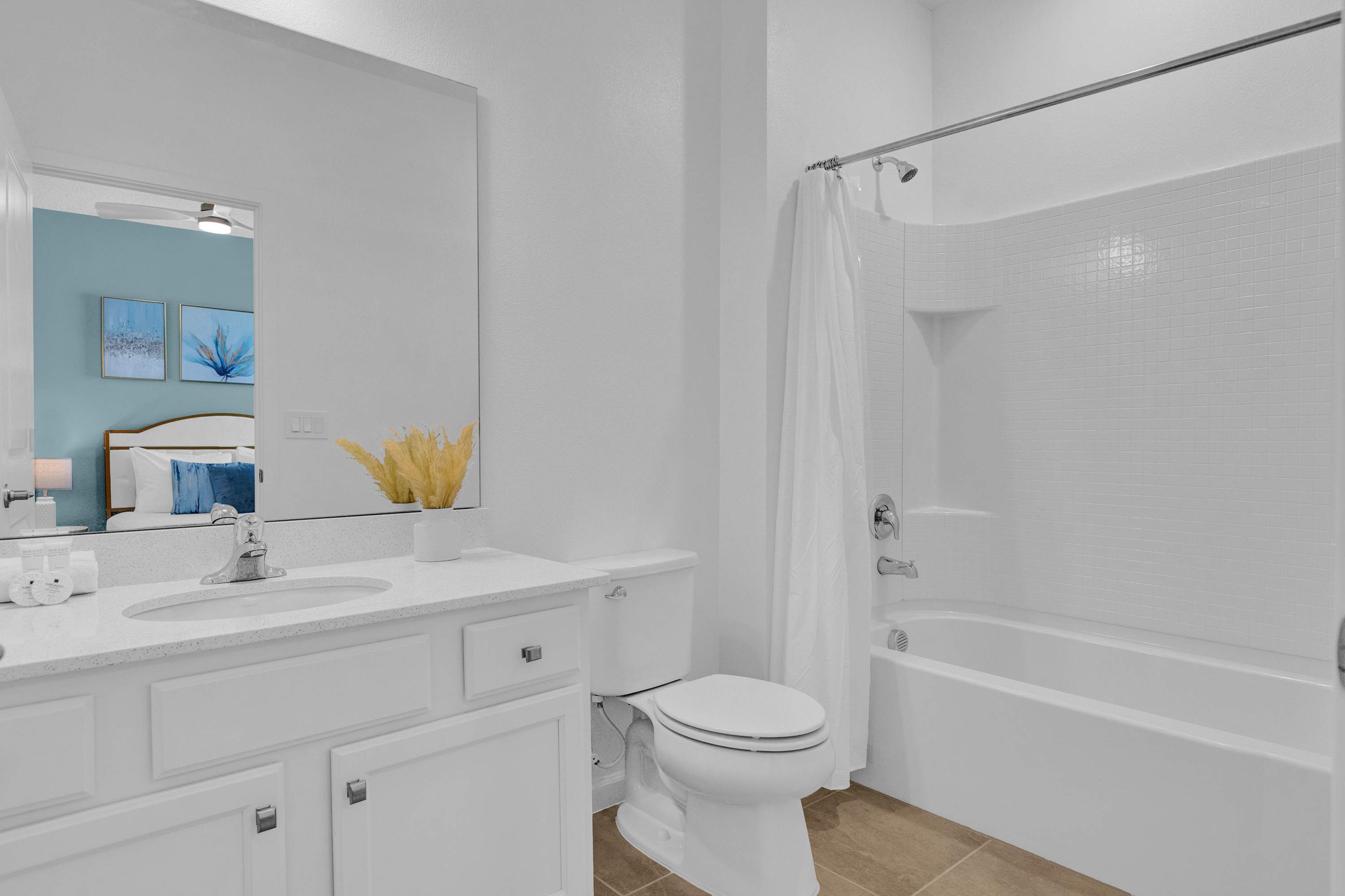 White bathroom with a single vanity, a toilet, and a bathtub with a white shower curtain. The mirror reflects the light blue wall and bed from an adjacent bedroom, and a small plant decorates the vanity.