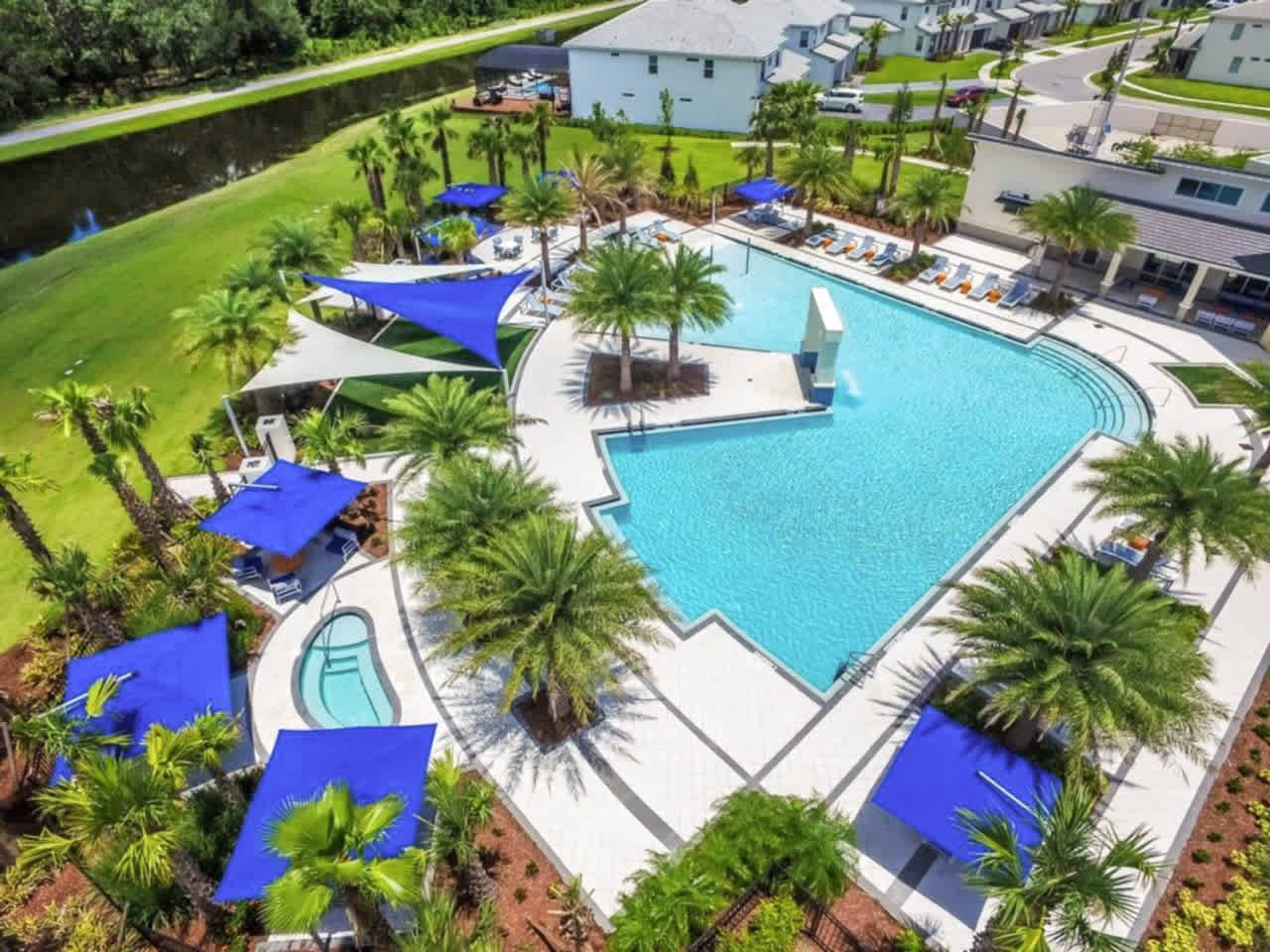 An aerial shot of a large, resort-style communal pool with multiple sections and a small hot tub, surrounded by numerous palm trees and blue shade structures. Lounge chairs are arranged around the pool deck, with a water feature visible.