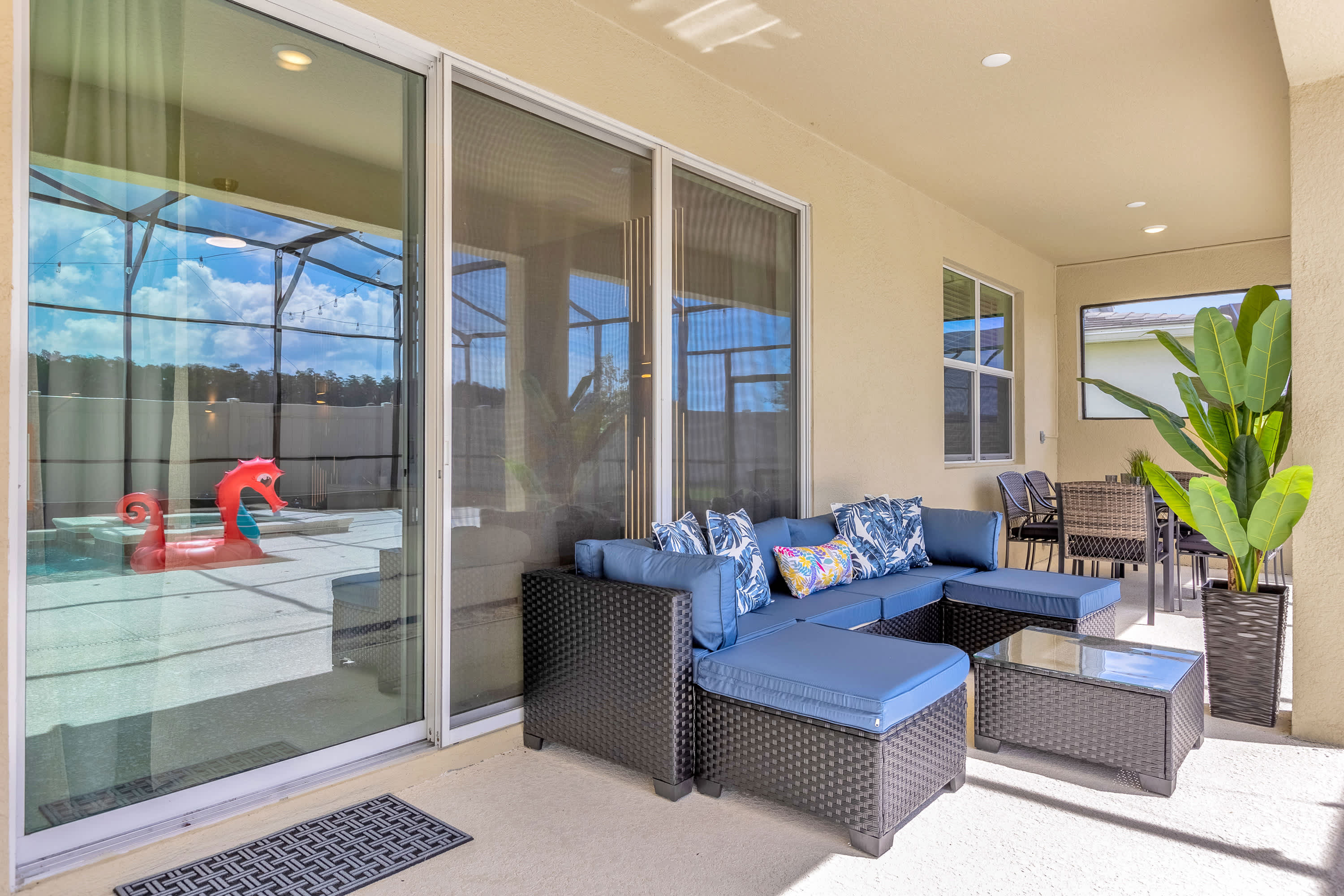 A comfortable outdoor lounge area on a covered patio, featuring a large blue sectional sofa with decorative pillows, a coffee table, and sliding glass doors leading indoors, with a view of the screened pool.