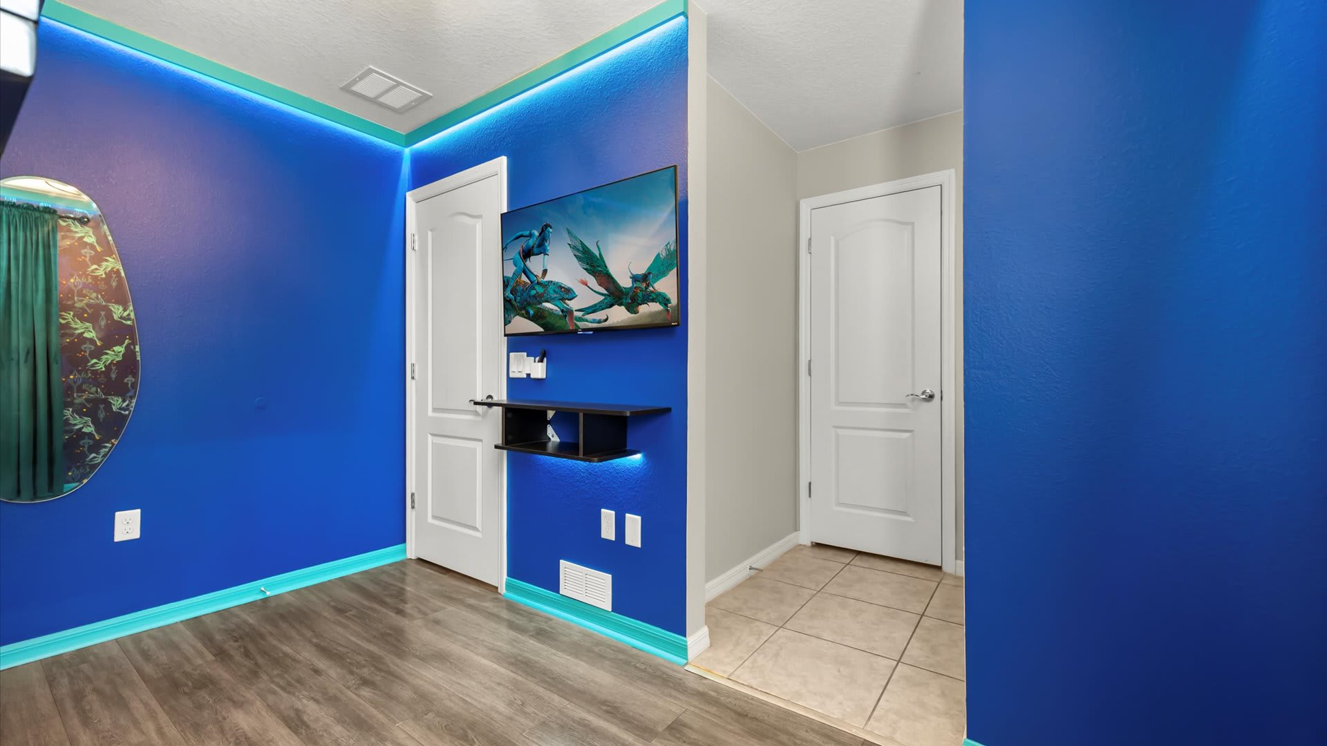 Themed bedroom with vibrant blue walls, a wall-mounted TV, and a floating black shelf. Teal trim, LED lighting, and a decorative mirror accent the room with wood-look flooring.