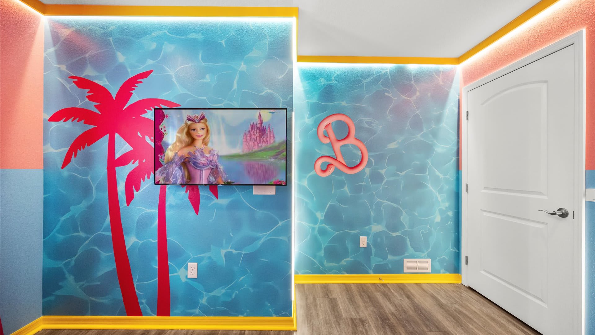 Barbie-themed room with a wall-mounted TV displaying a Barbie movie, set against a blue wall with a palm tree graphic and water ripple effect. A large pink 'B' symbol adorns an adjacent wall.