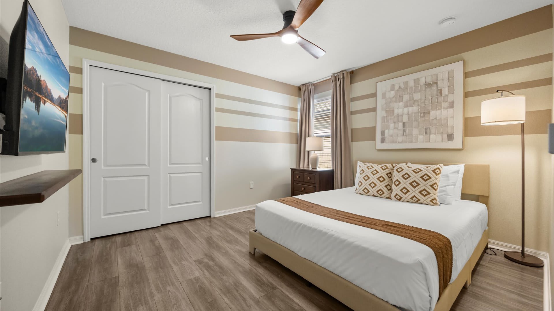 A cozy bedroom features a queen-sized bed with white bedding and a brown runner, a dark wood nightstand, and a large abstract artwork above the bed. The walls are decorated with horizontal stripes in beige and white, and a wooden ceiling fan hangs overhead.