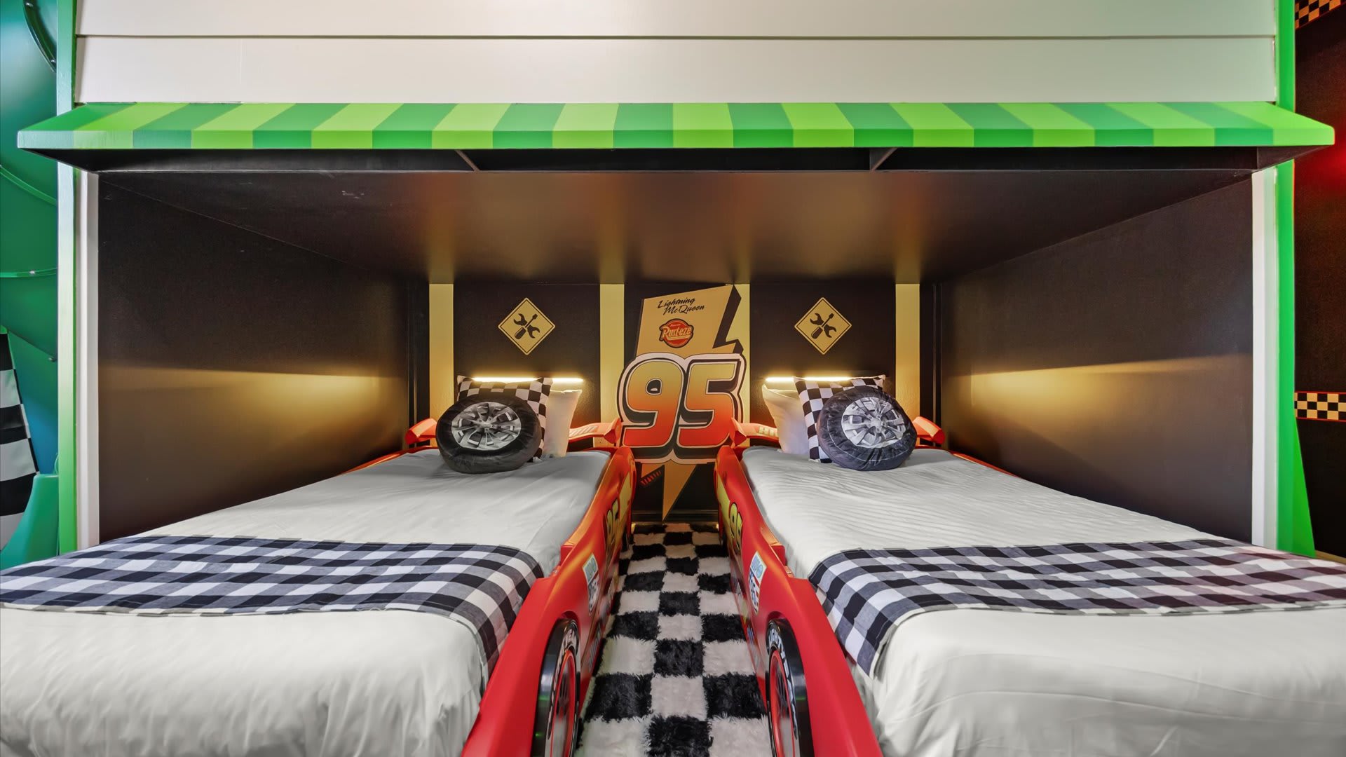 A close-up view of two red race car beds with checkerboard bedding, nestled under a bunk bed structure adorned with a '95' logo in the Cars-themed room.