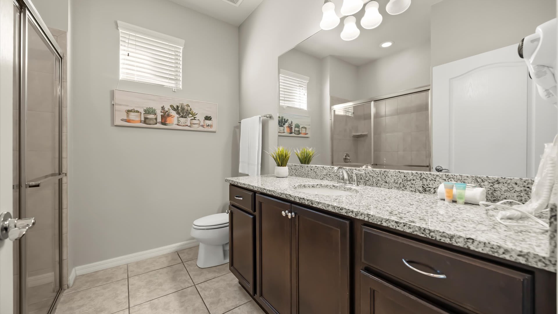A bright bathroom features a dark wood vanity with a granite countertop and single sink, a large mirror, and a walk-in shower with a glass door. Light gray walls, tiled floors, and a window with blinds complete the functional space.