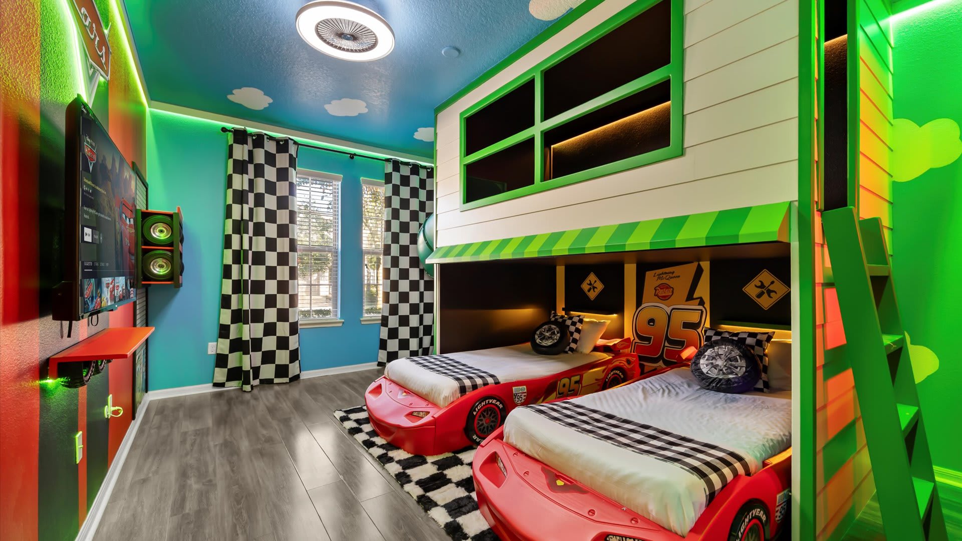 A lively Cars-themed bedroom featuring two red race car beds, a built-in bunk bed structure, checkerboard curtains, and a wall-mounted TV with speakers.