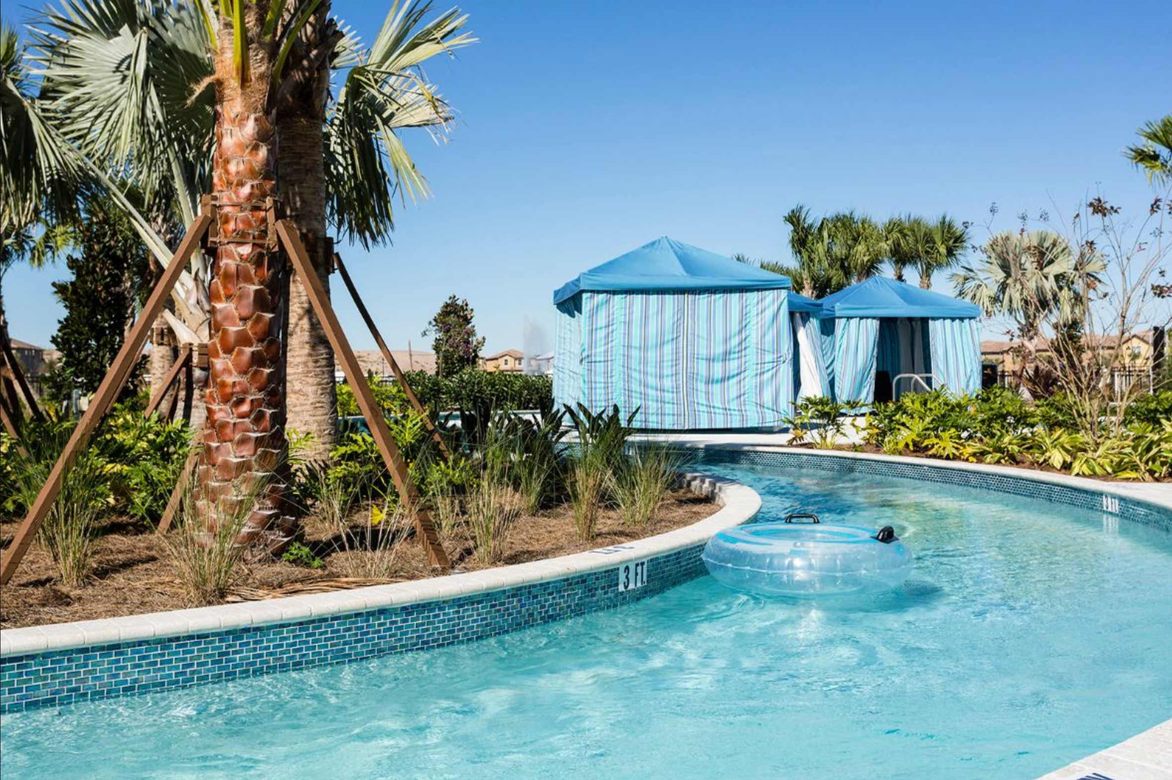 A serene lazy river with clear blue water and blue mosaic tile edging, featuring an inflatable tube floating gently. Lush palm trees and blue-striped cabanas line the banks, creating a relaxing resort atmosphere.
