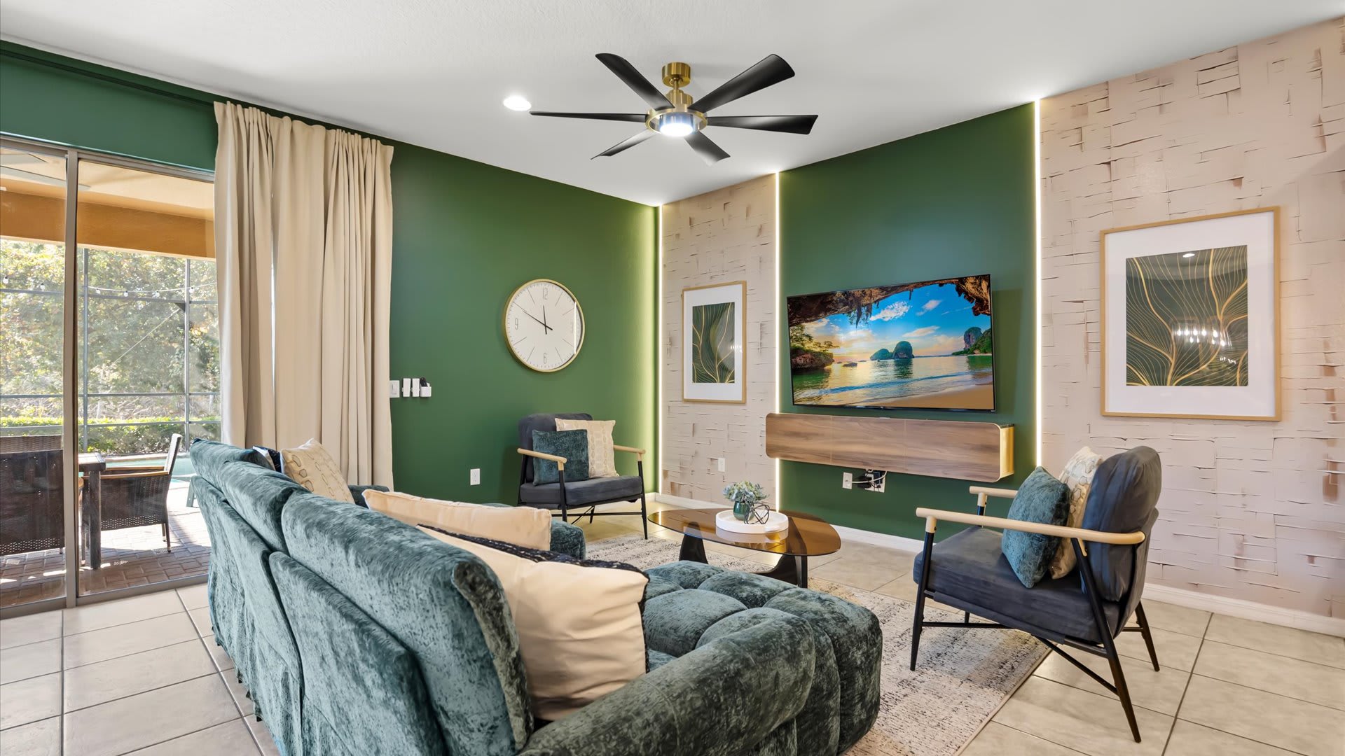 A modern living room with dark green accent walls, a plush teal sofa, two armchairs, and a wall-mounted TV. The room features light-colored tile flooring, a ceiling fan, and a large sliding glass door to the patio.