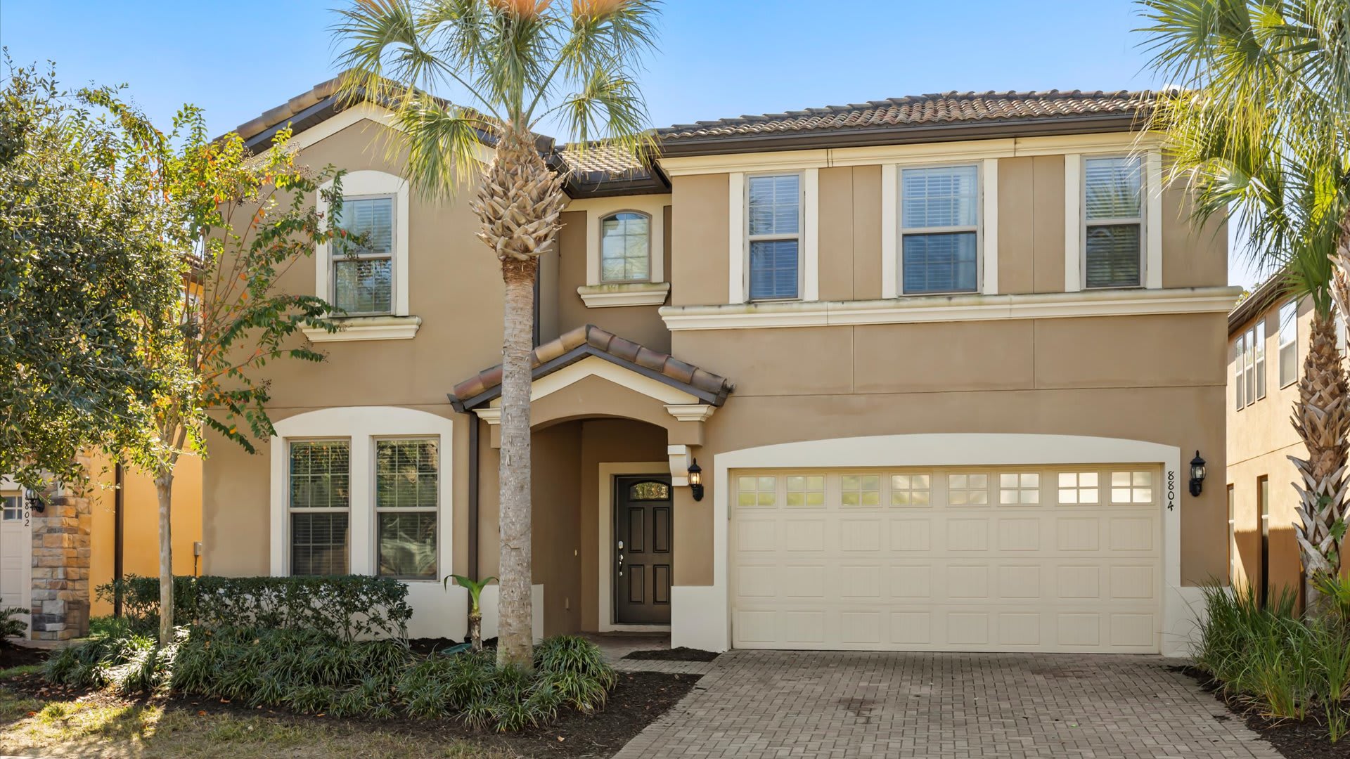 Grand two-story house with a beige stucco exterior, dark brown roof tiles, and a paved driveway leading to a two-car garage. Lush palm trees and green landscaping frame the entrance.