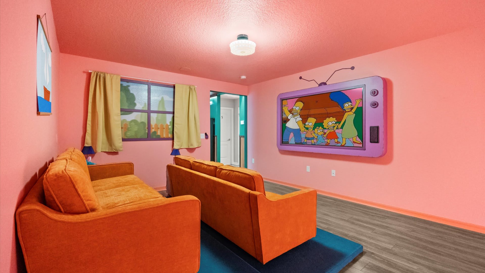 A playful media room with bright pink walls, two orange sofas, and a large cartoon-style television displaying The Simpsons. The room features wood-look flooring and a window with light curtains.