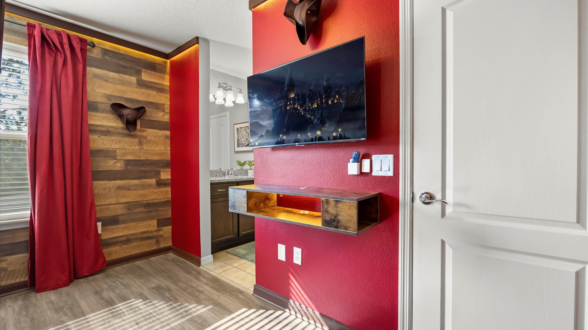 A red accent wall with a wall-mounted TV and a floating wooden shelf, adjacent to a wood-paneled wall and a white door leading to a bathroom, within the pirate-themed room.