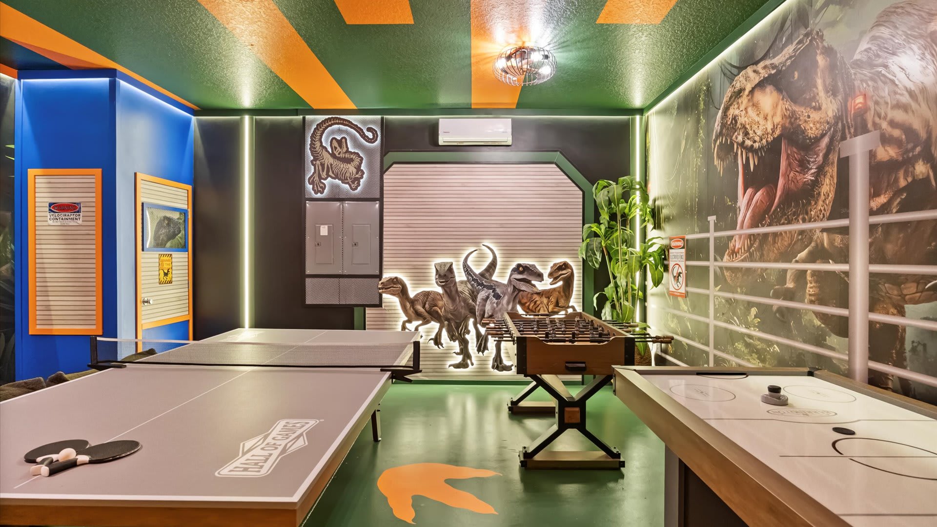 A Jurassic Park-themed game room with a ping pong table, foosball table, and air hockey table. The room features dark green walls with dinosaur murals, orange and green striped ceiling, and glowing dinosaur cutouts.