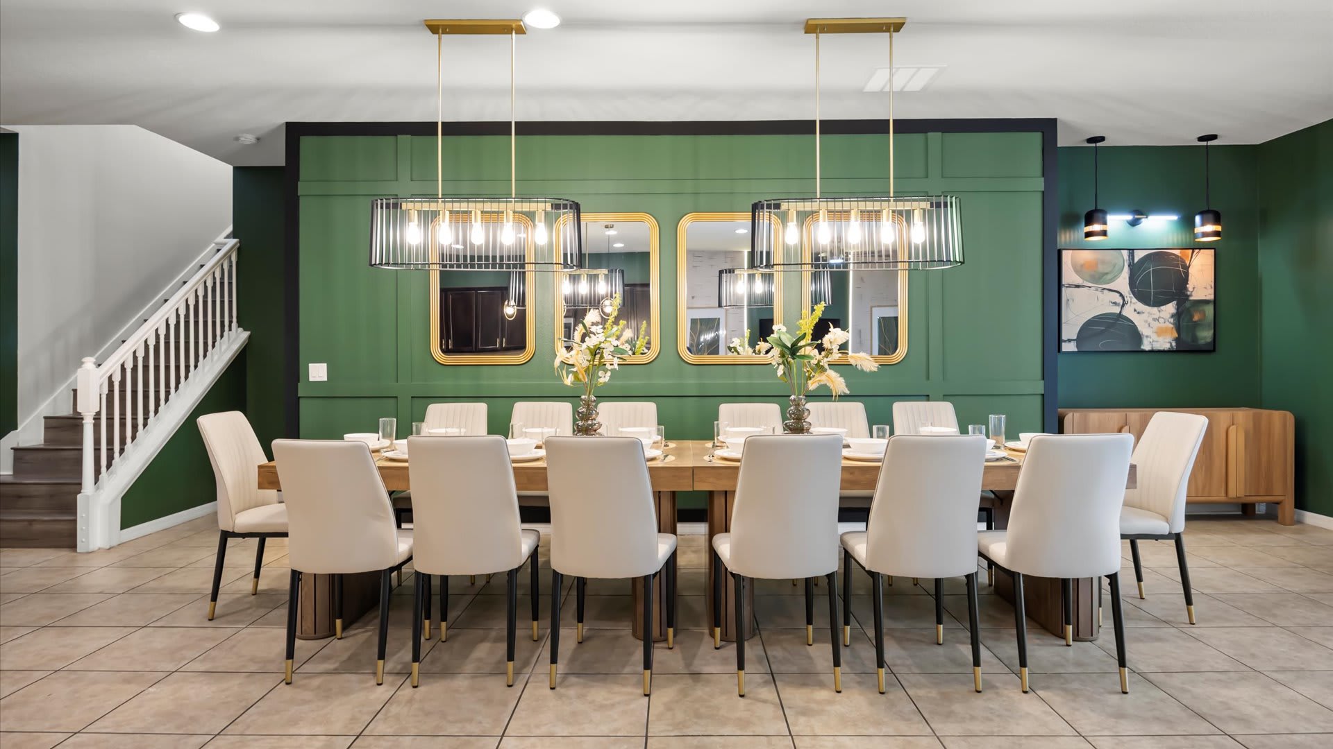 A grand dining room with a long wooden table and twelve chairs, flanked by a dark green paneled accent wall with mirrors and modern chandeliers. A staircase with white railings is visible in the background, all on light tile flooring.