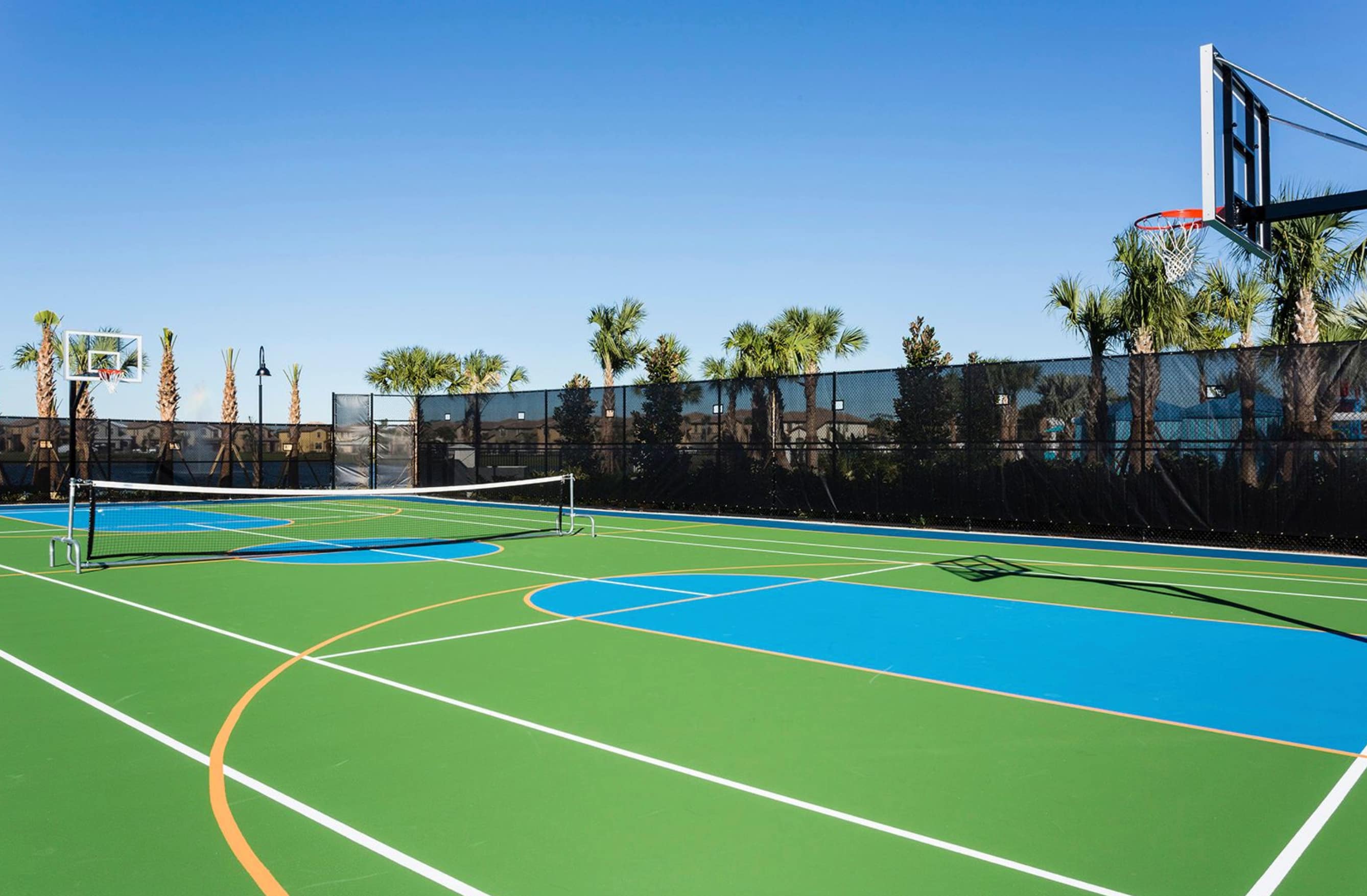 A bright outdoor multi-sport court with green and blue surfaces, marked for basketball and volleyball, features two basketball hoops and a net, enclosed by a fence with palm trees in the background.
