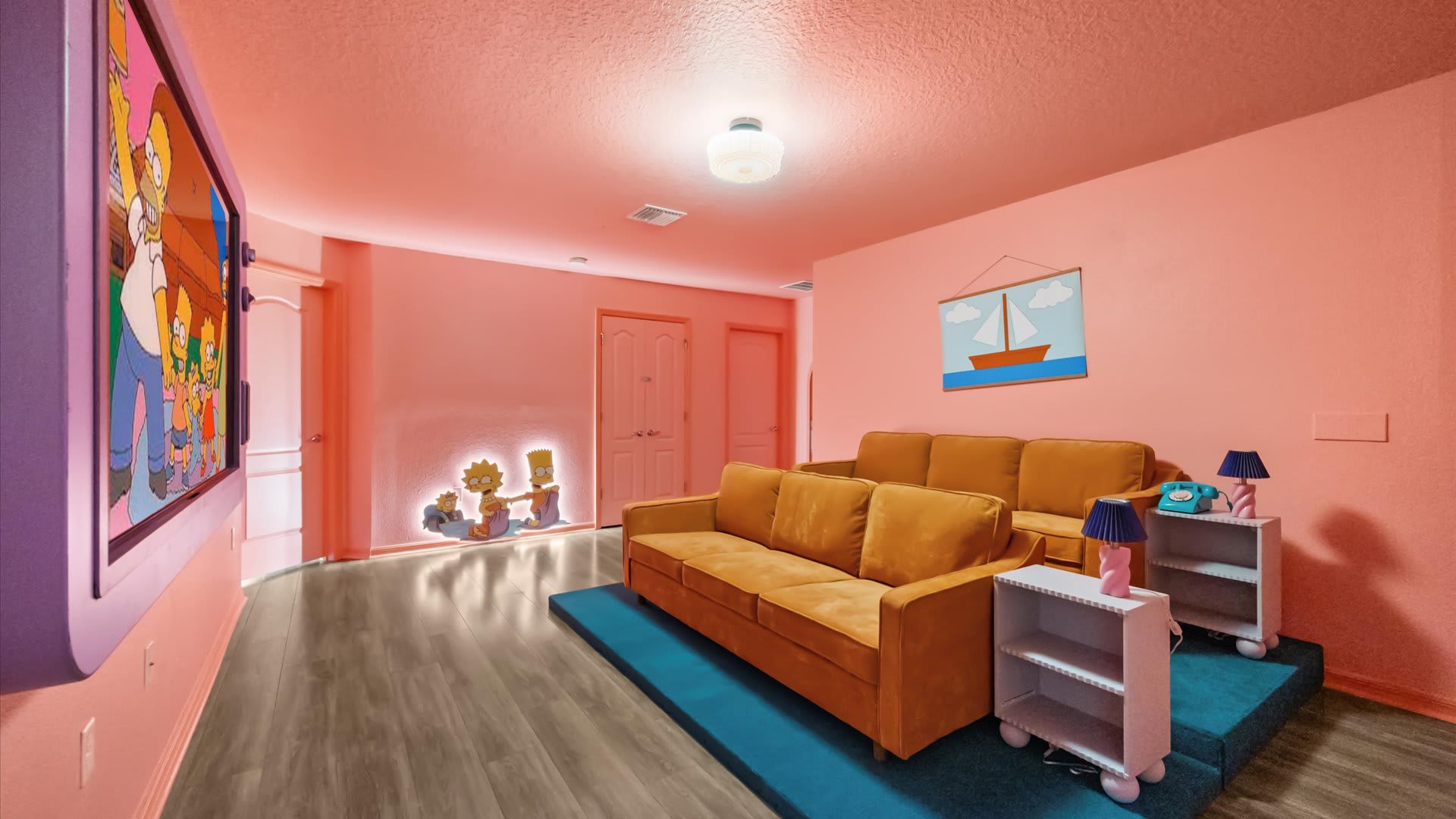 A pink-walled media room, themed after The Simpsons, features two rows of mustard-yellow sofas on a raised platform with a blue carpet. A large TV screen displays a cartoon, and character cutouts illuminate the wall.