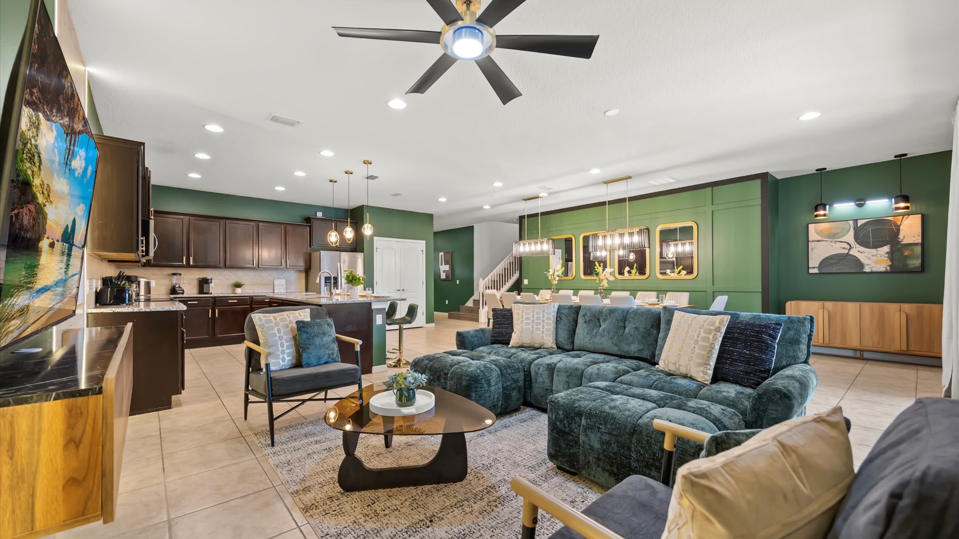 A spacious open-concept living area features a dark green sectional sofa, two armchairs, and a glass coffee table on a patterned rug, with a dark wood kitchen and dining area visible in the background. The walls are painted a deep green, and a large ceiling fan hangs overhead.