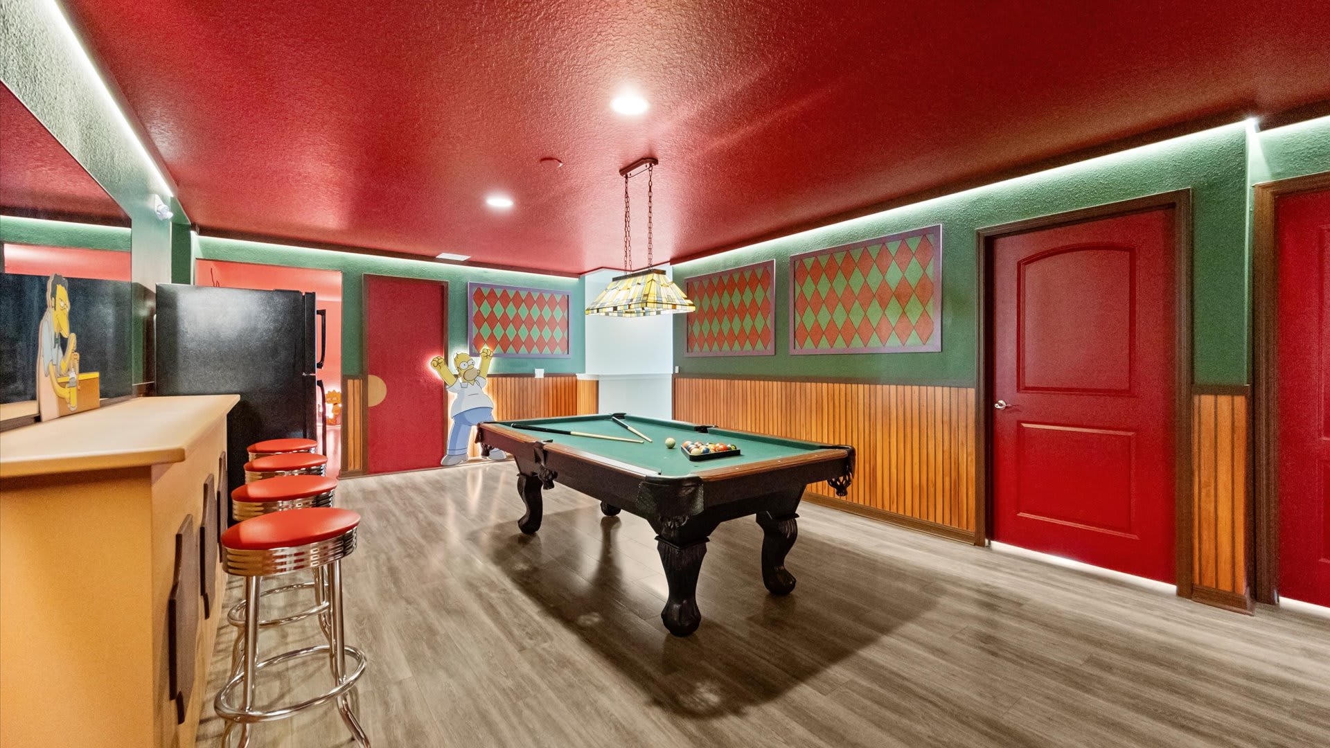 A vibrant game room featuring a pool table, a bar with red stools, and Simpsons-themed decor with red ceilings and green walls. The room has wood-look flooring and recessed lighting.