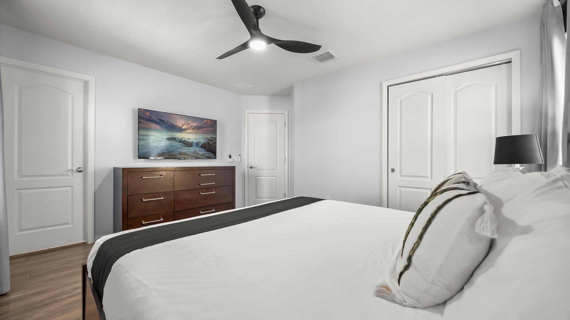 A bright bedroom features a large bed with white bedding and a black runner, a dark wood dresser with a mounted TV, and a black ceiling fan. White doors and light gray walls complete the modern look.