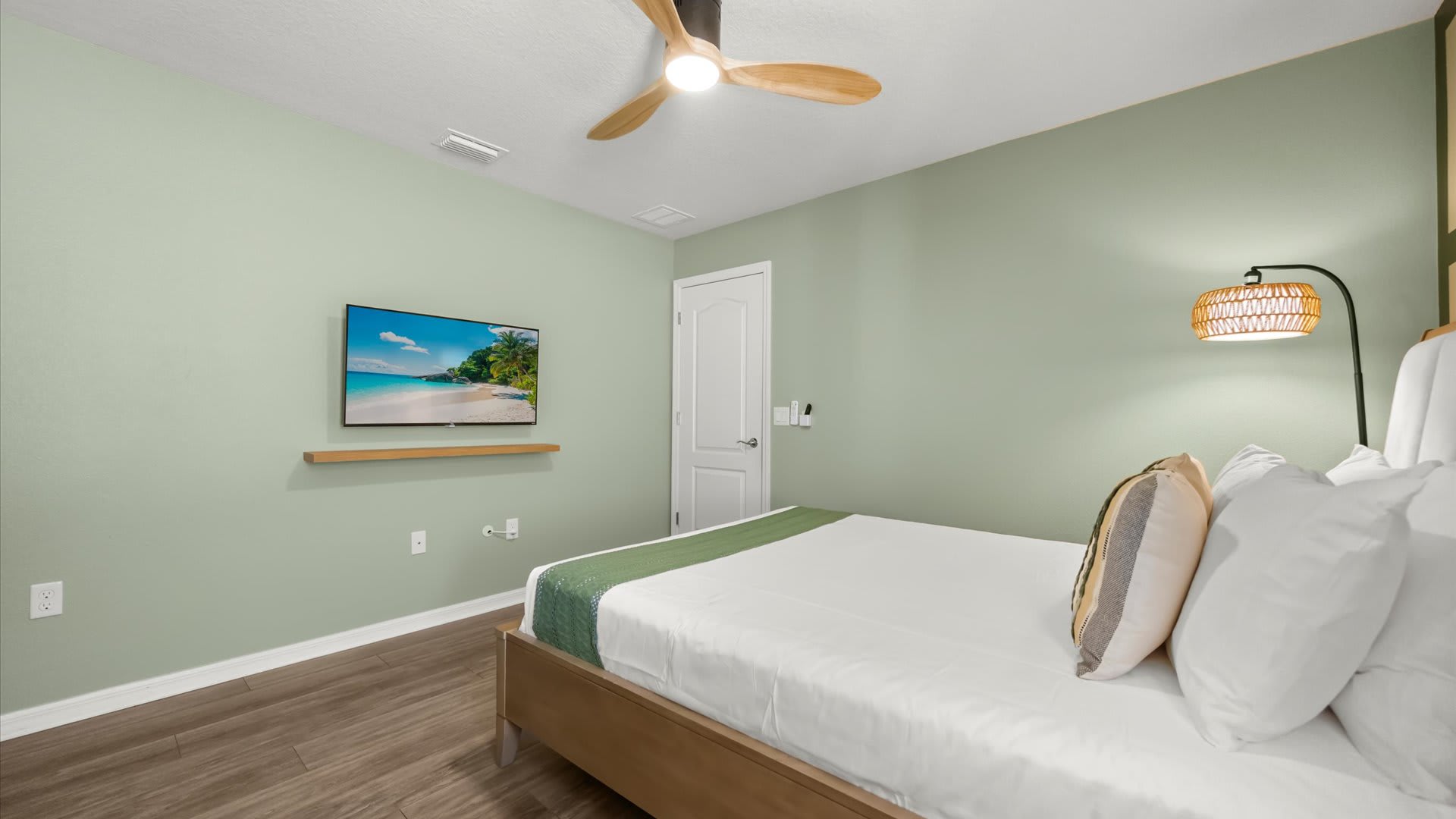 This bedroom features a queen-sized bed with white bedding and a green runner, a wall-mounted TV on a floating shelf, and a unique floor lamp. The light green walls and wood-look flooring create a serene and inviting atmosphere.