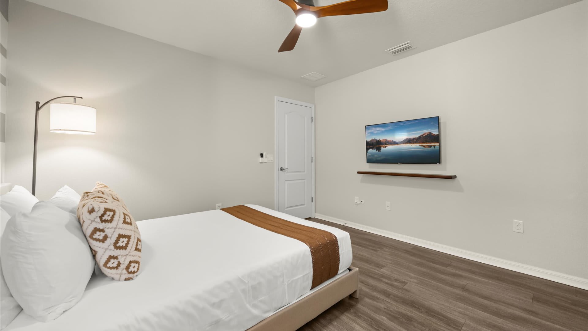 This bedroom features a queen-sized bed with white bedding and a brown runner, a wall-mounted TV on a floating shelf, and a modern floor lamp. The light gray walls and wood-look flooring create a minimalist and comfortable space.