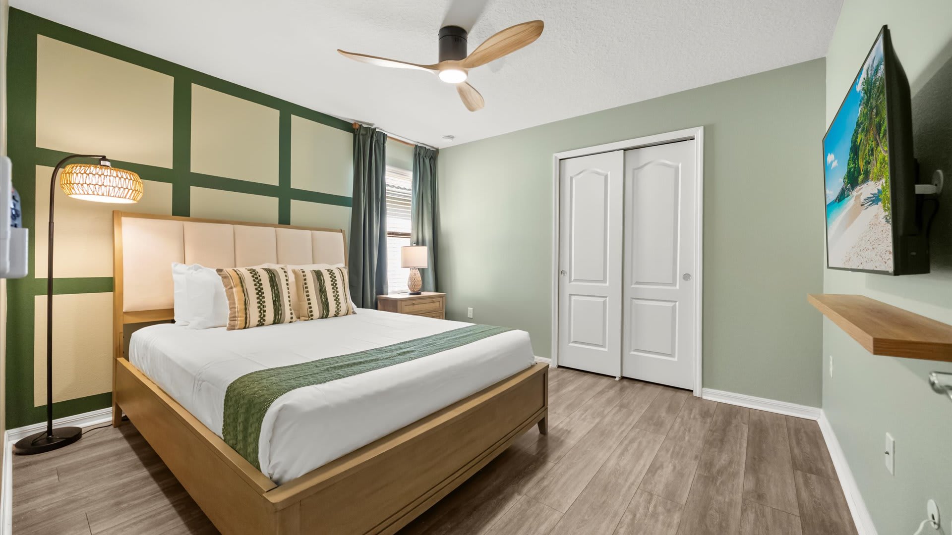 A vibrant bedroom features a queen-sized bed with a light wood frame and upholstered headboard, green and white accent walls, and a wooden ceiling fan. A wall-mounted TV and a unique floor lamp add to the modern decor.