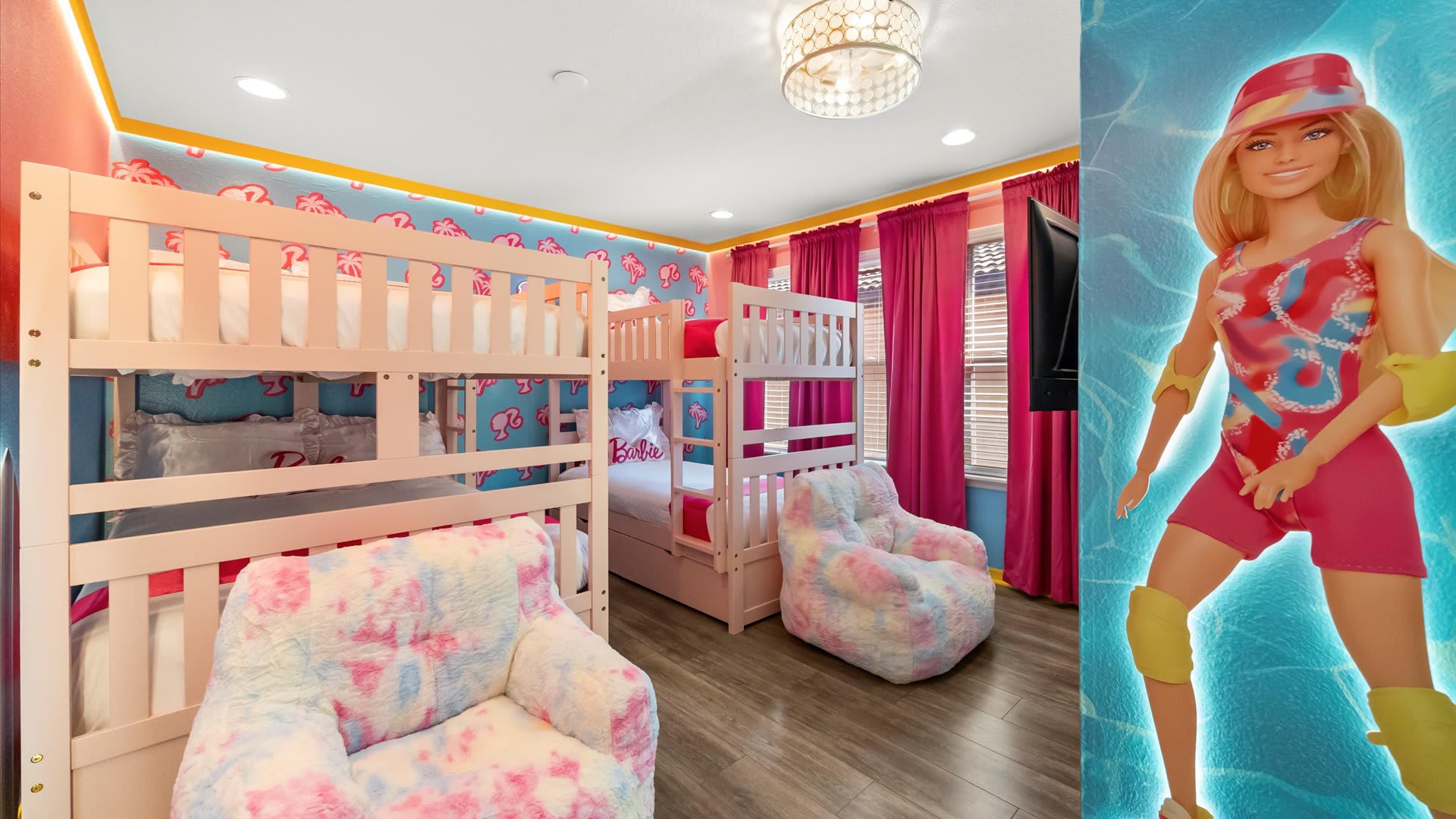 Barbie-themed bedroom featuring two white bunk beds with pink accents, a large Barbie graphic on the wall, and two tie-dye bean bag chairs. The room has wood-look flooring and pink curtains.