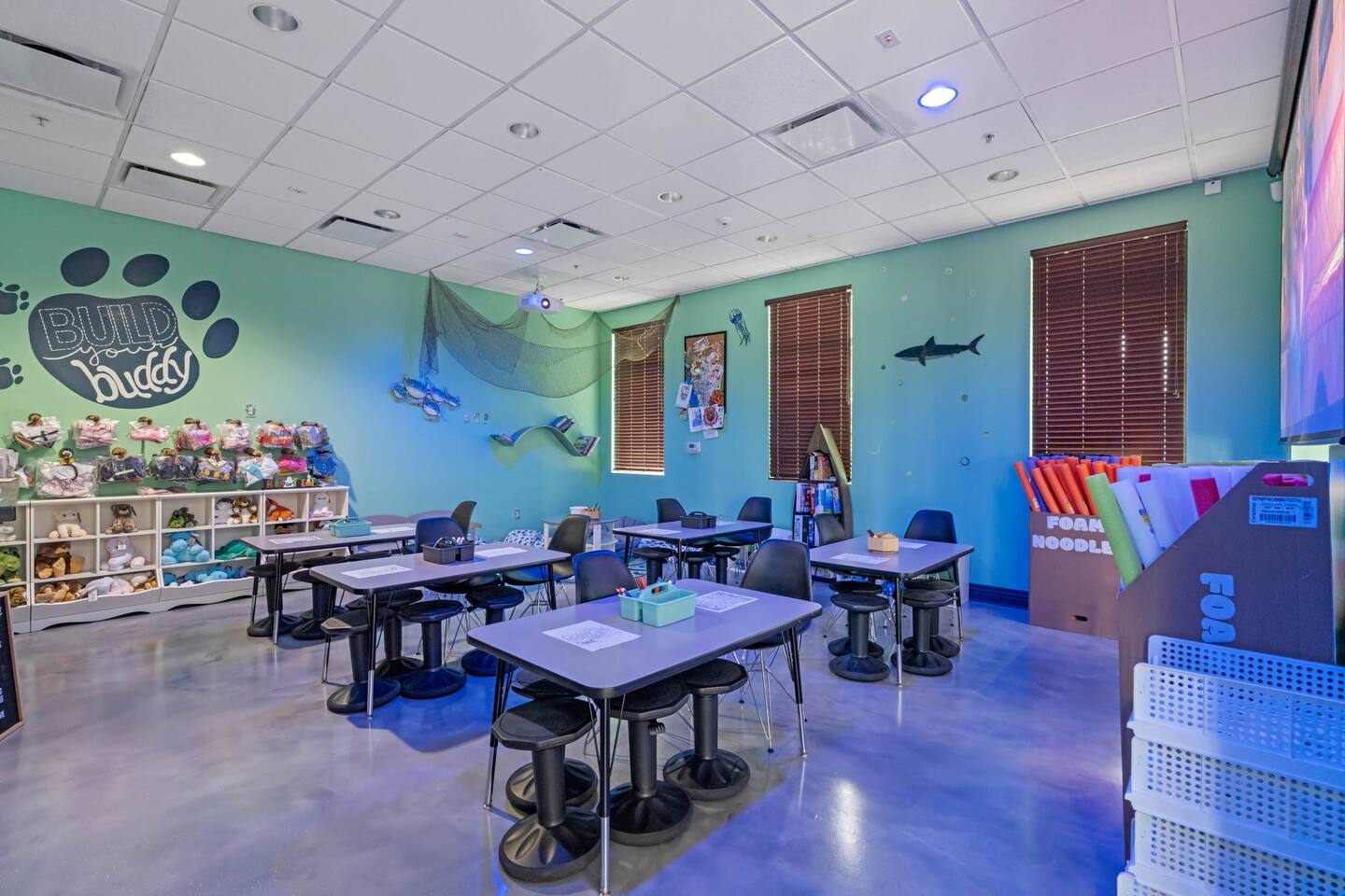 A colorful kids' activity room features light blue walls, several tables and stools, shelves filled with stuffed animals, and ocean-themed decorations like a shark cutout and fishing net.