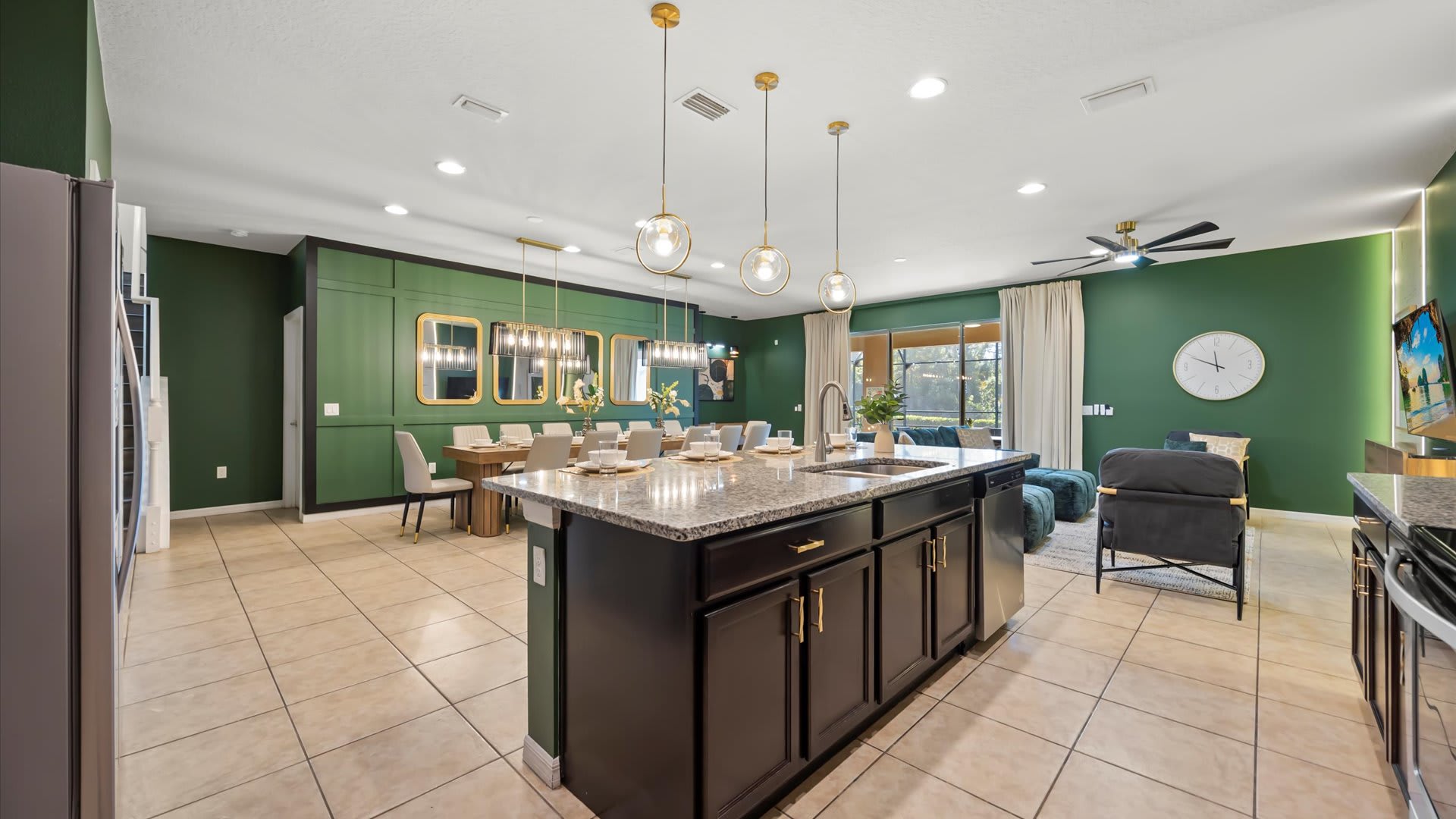 An open-concept living space showcasing a modern kitchen with an island, a formal dining area, and a cozy living room. The cohesive design features dark green accent walls, tile flooring, and contemporary lighting fixtures throughout.