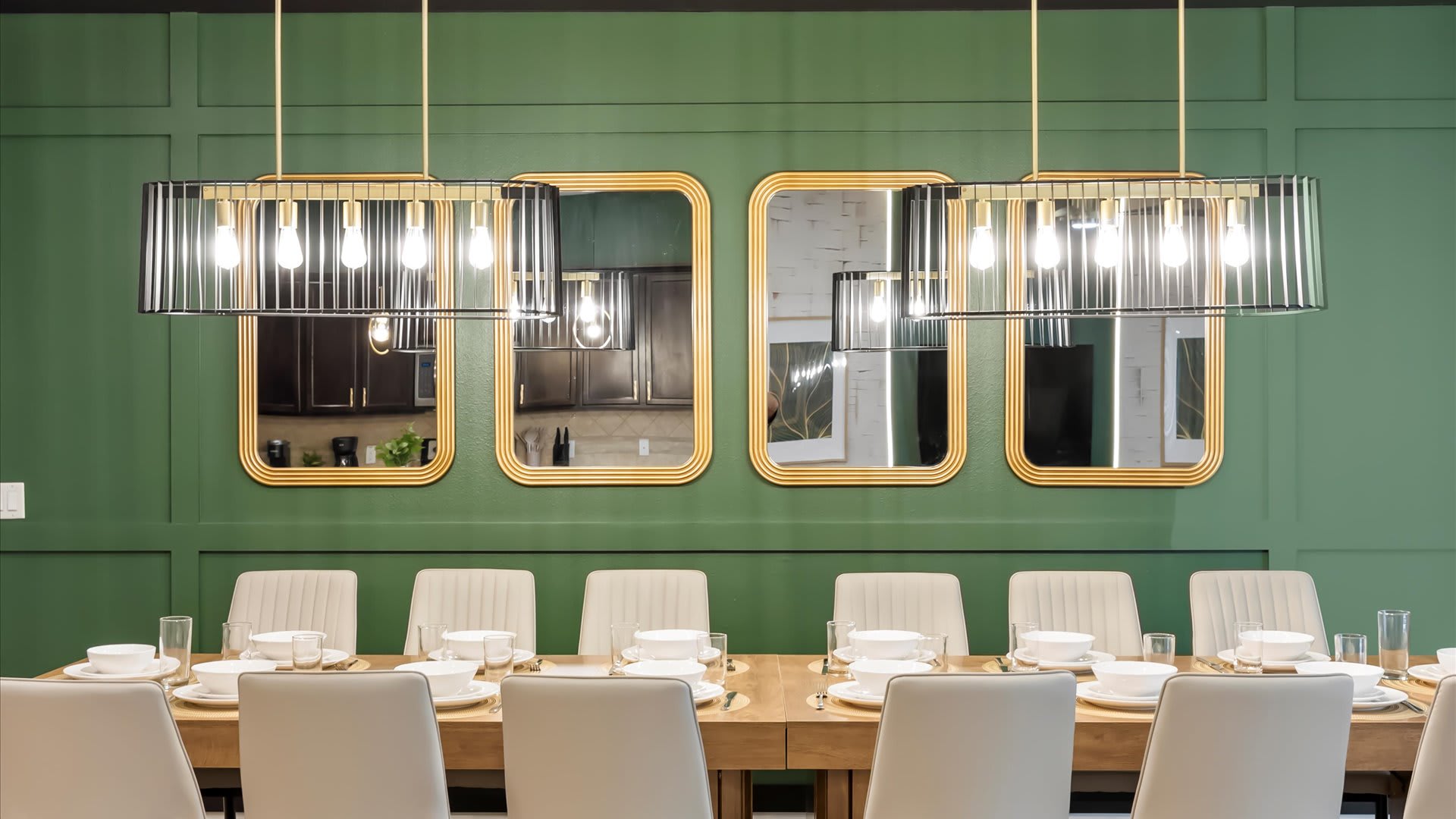 A close-up view of the elegant dining table set for twelve, featuring white place settings and floral centerpieces. The dark green paneled accent wall is adorned with two modern chandeliers and gold-framed mirrors.