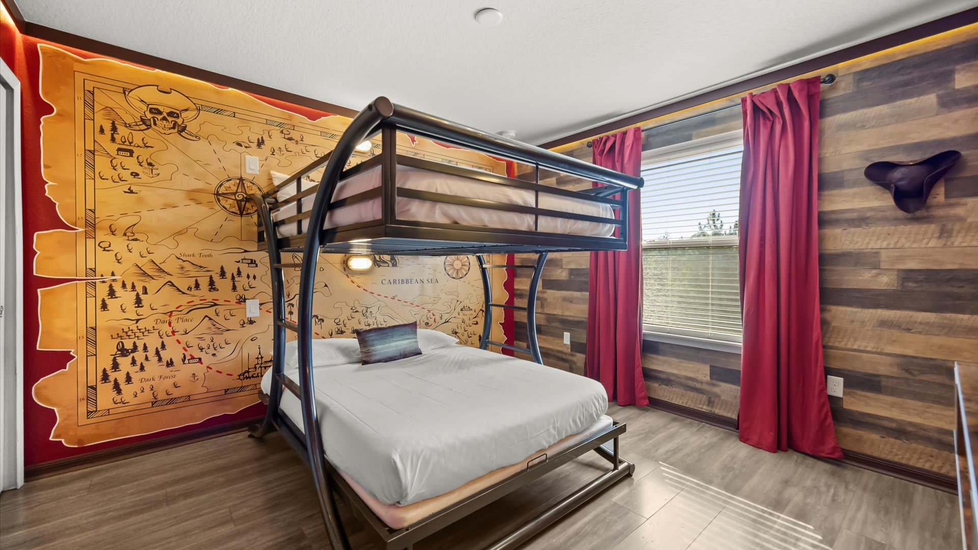 A pirate-themed bedroom featuring a sturdy metal bunk bed (full over full) against a large pirate map mural, complemented by wood-paneled accent walls and red curtains.