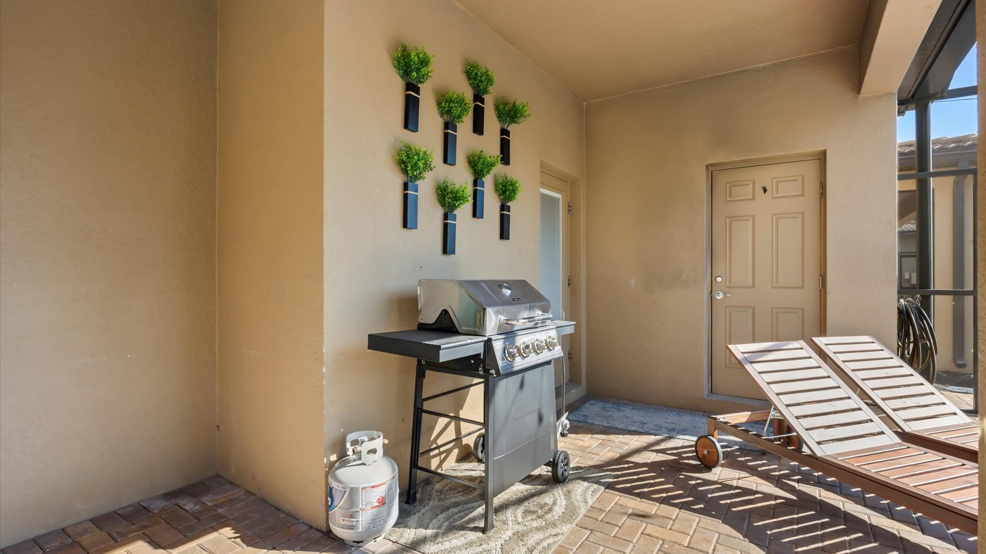 A dedicated outdoor grilling area on the covered patio features a large gas BBQ grill with a propane tank, next to two wooden lounge chairs. Decorative wall planters add a touch of greenery to the beige wall.