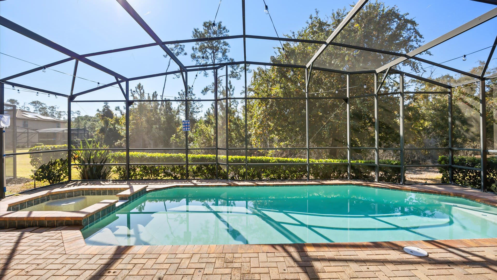A serene view of the screened-in swimming pool and hot tub, surrounded by a brick paver deck and backed by dense green foliage and tall trees. The clear blue water reflects the bright sky.