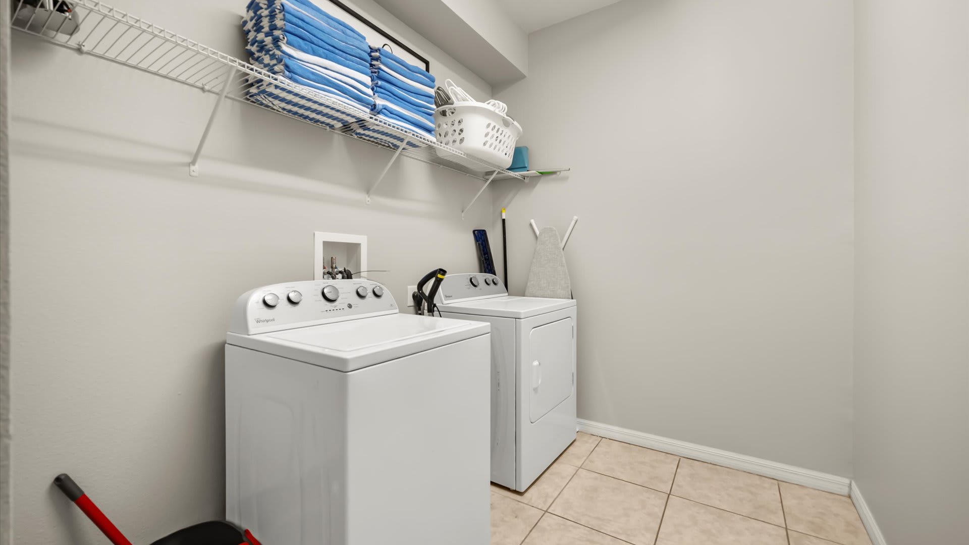 Functional laundry room featuring a white top-load washing machine and a white dryer, with a wire shelf above holding neatly folded blue towels. The room has light gray walls and tiled flooring.