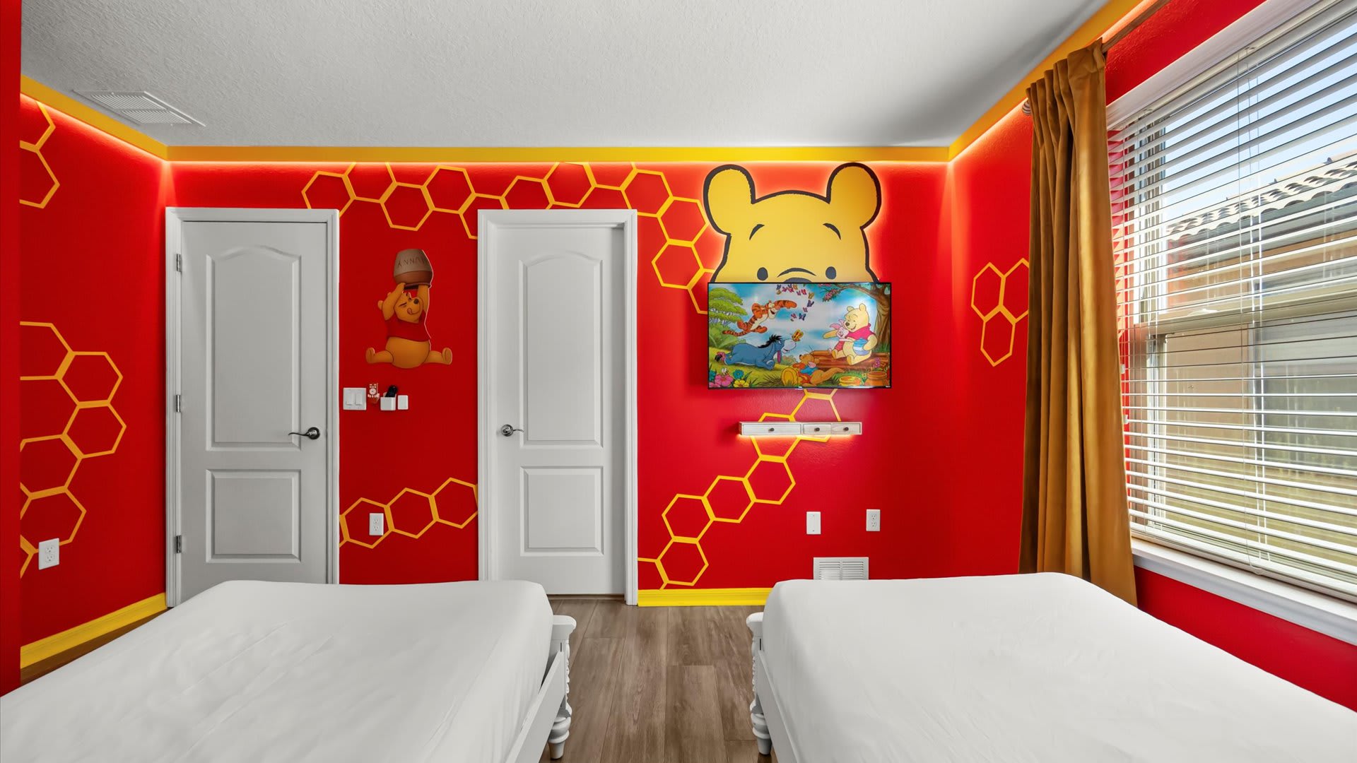 A vibrant Winnie the Pooh themed bedroom with bright red walls, yellow honeycomb patterns, two single beds, and a wall-mounted TV under a large Pooh bear headboard.